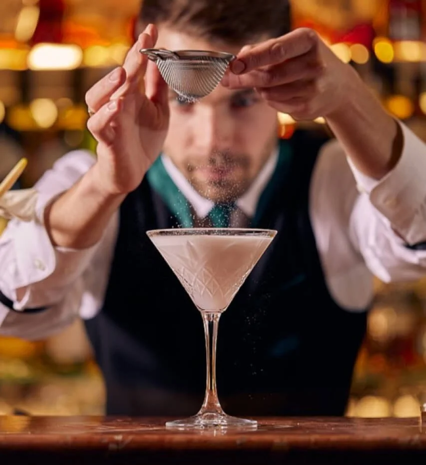 Bartender pouring a cloudy pink cocktail through a small strainer into a martini glass.