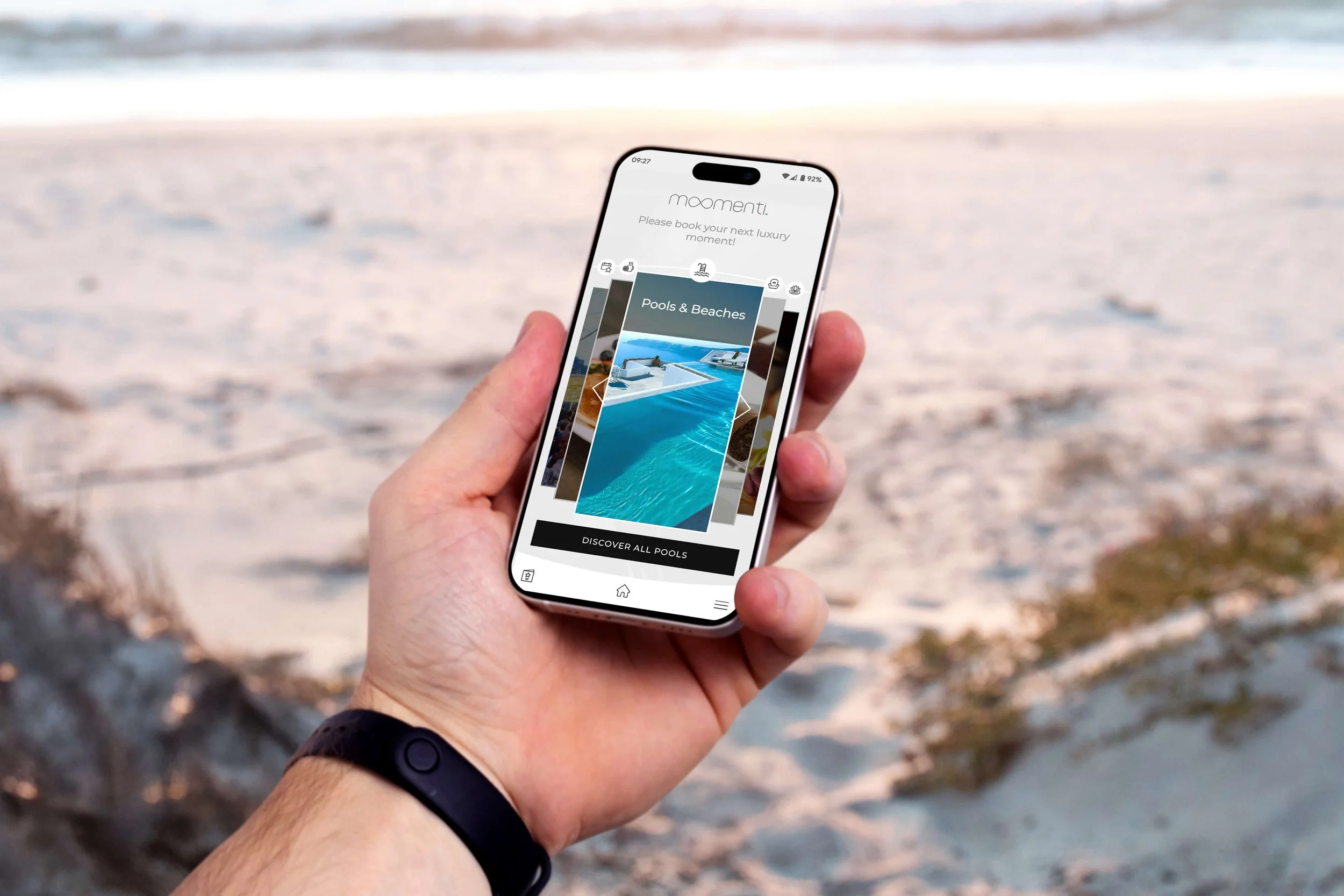 Person holding a smartphone with a travel app open, showing options for pools and beaches, against a sandy beach background with ocean waves.