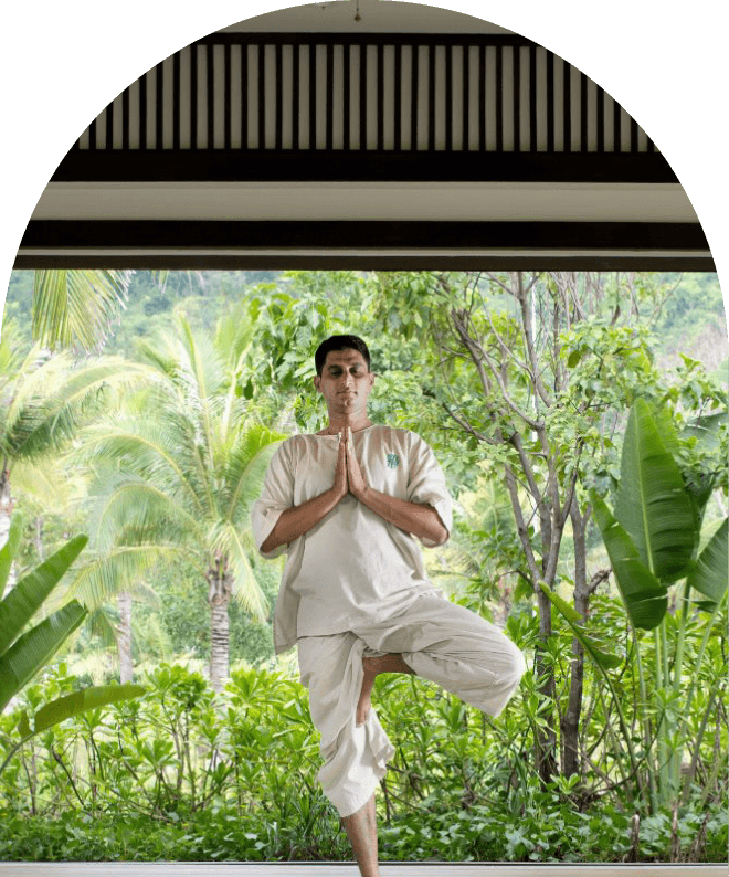 A man practicing yoga outdoors in a garden, standing on one leg with hands in prayer position, surrounded by lush green tropical plants and trees.