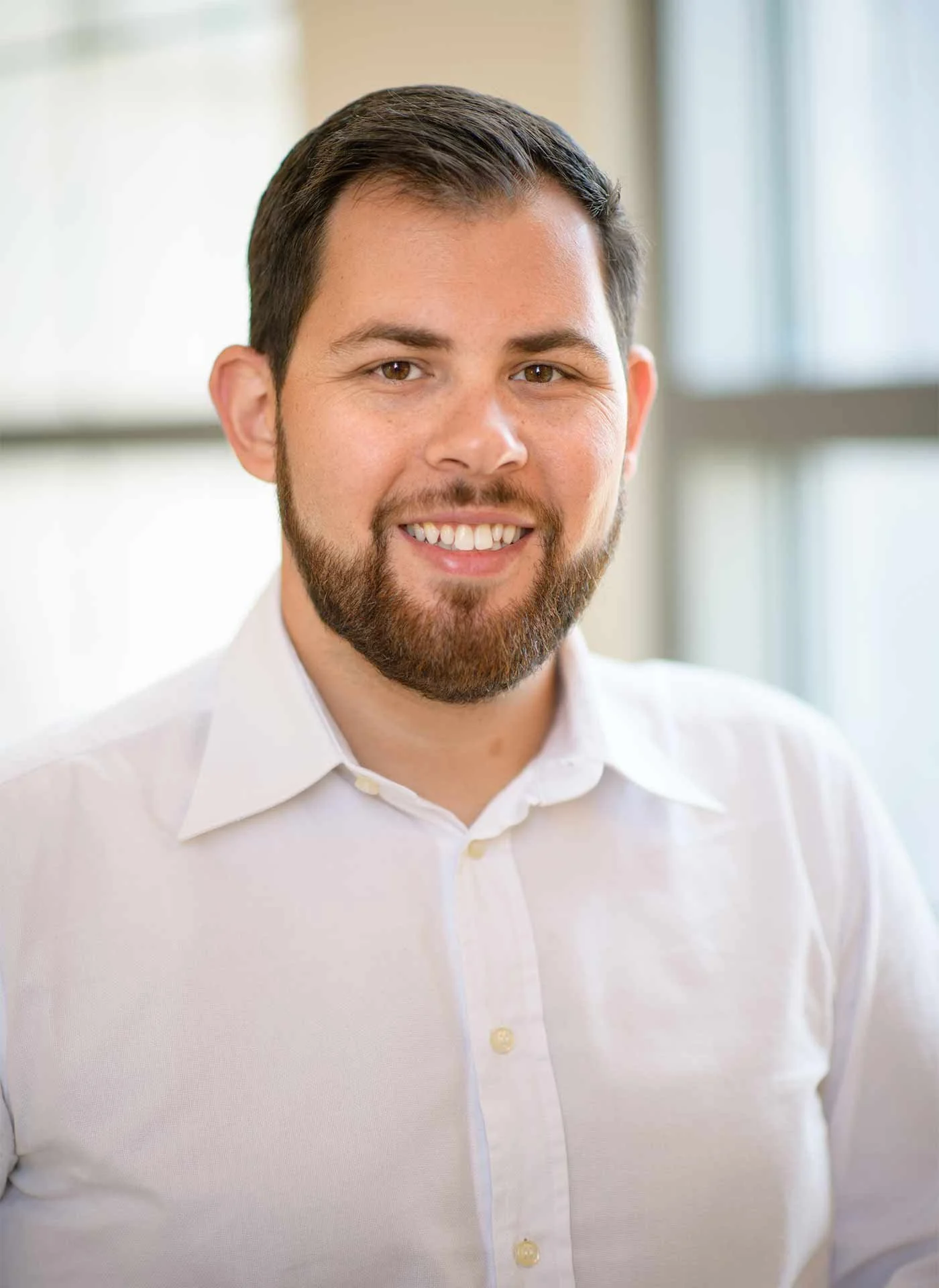 A smiling man with dark hair and a beard, wearing a white shirt, standing in front of a window in a bright indoor setting.
