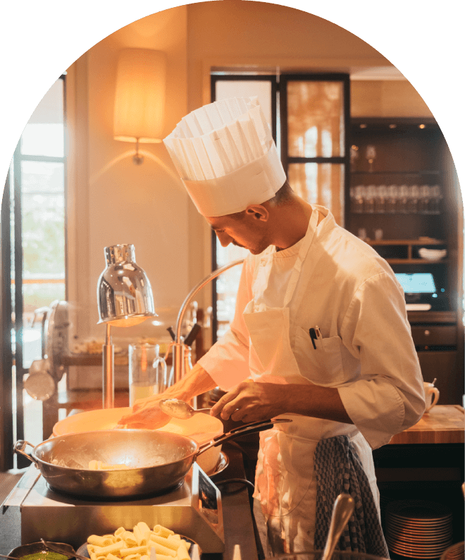Chef cooking in a kitchen with natural light, wearing a tall white chef's hat and apron, stirring food in a pan.