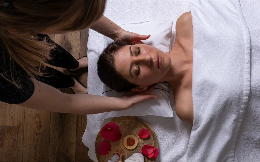 A woman receiving a head massage during a spa treatment, lying on a bed with a towel, surrounded by rose petals and spa products.