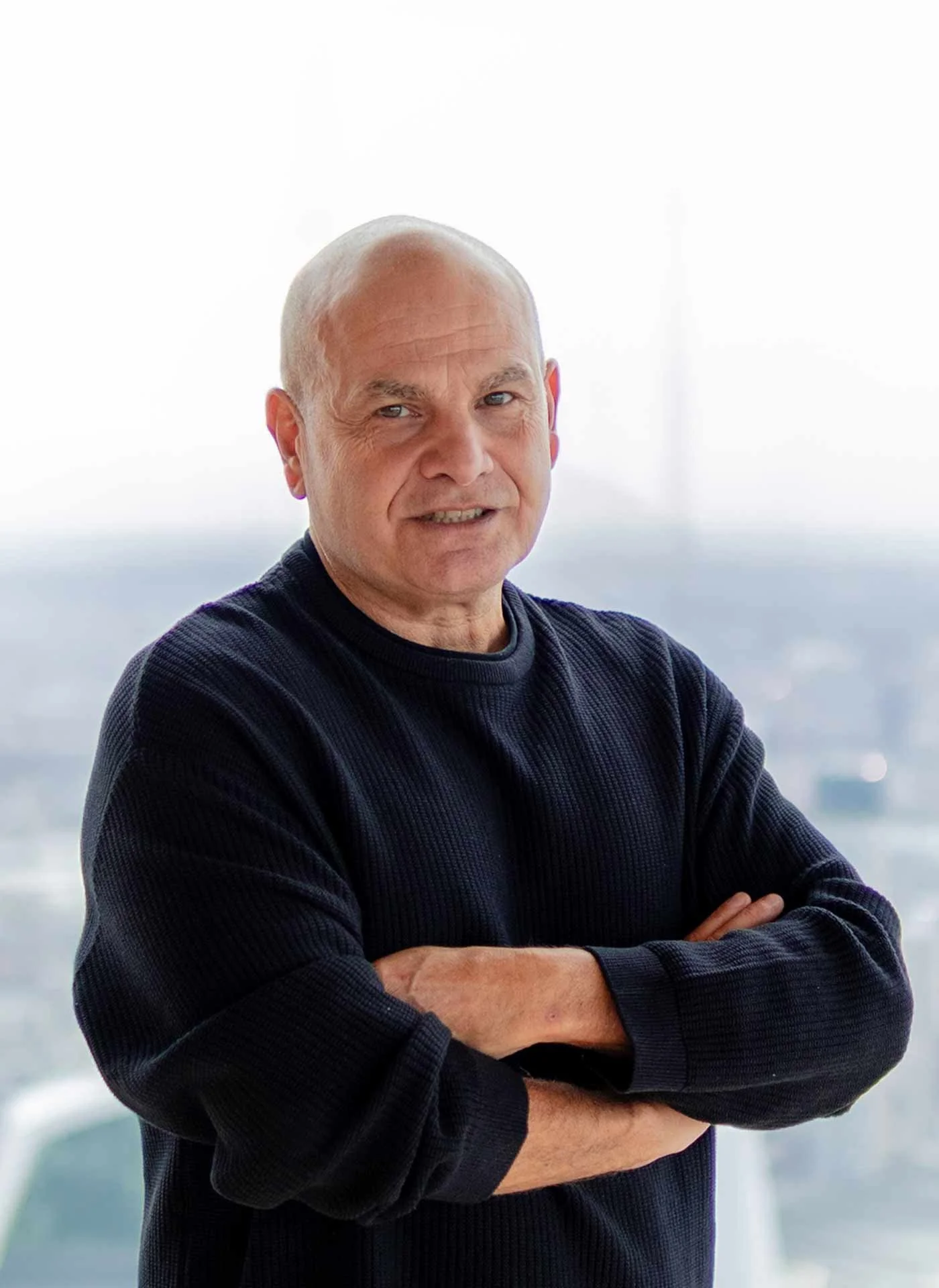 An older man with a bald head and light complexion standing with arms crossed indoors. He is wearing a black sweater and is in front of a window with a blurred cityscape background.
