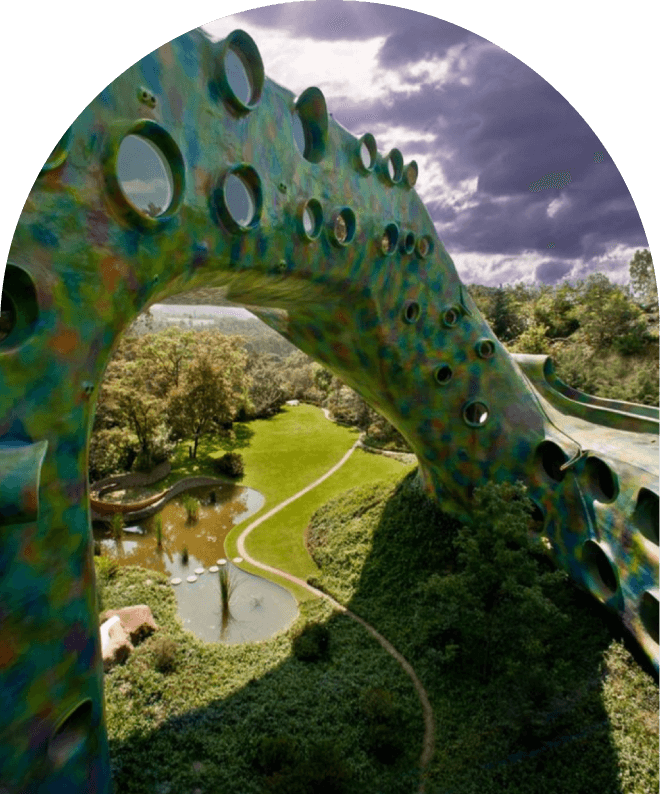 Bird's eye view of a large, colorful playground structure that resembles a bridge, over a park with trees, a pond, walking paths, and a cloudy sky in the background.