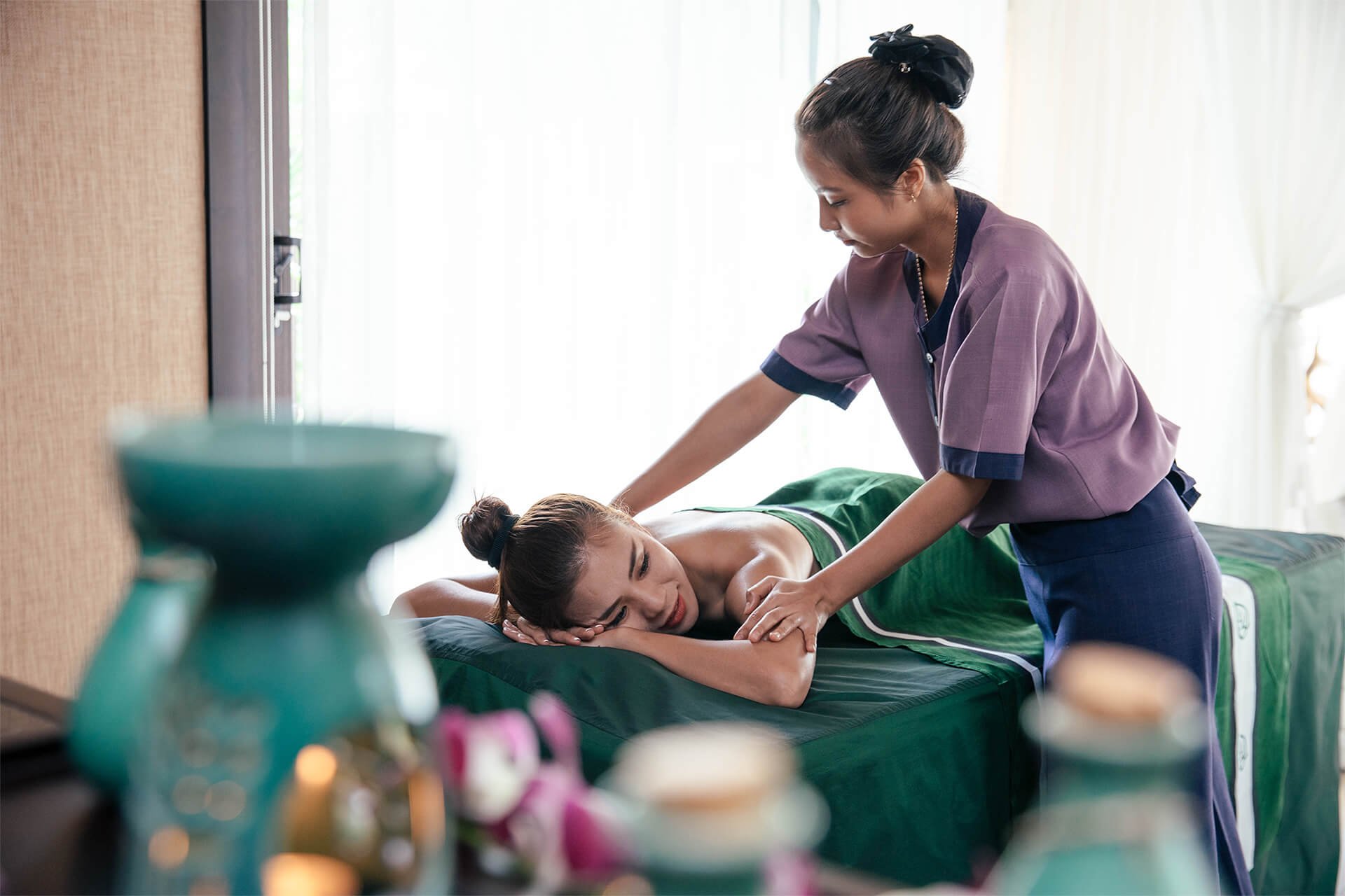 A massage therapist giving a massage to a woman lying face down on a massage table in a bright room.