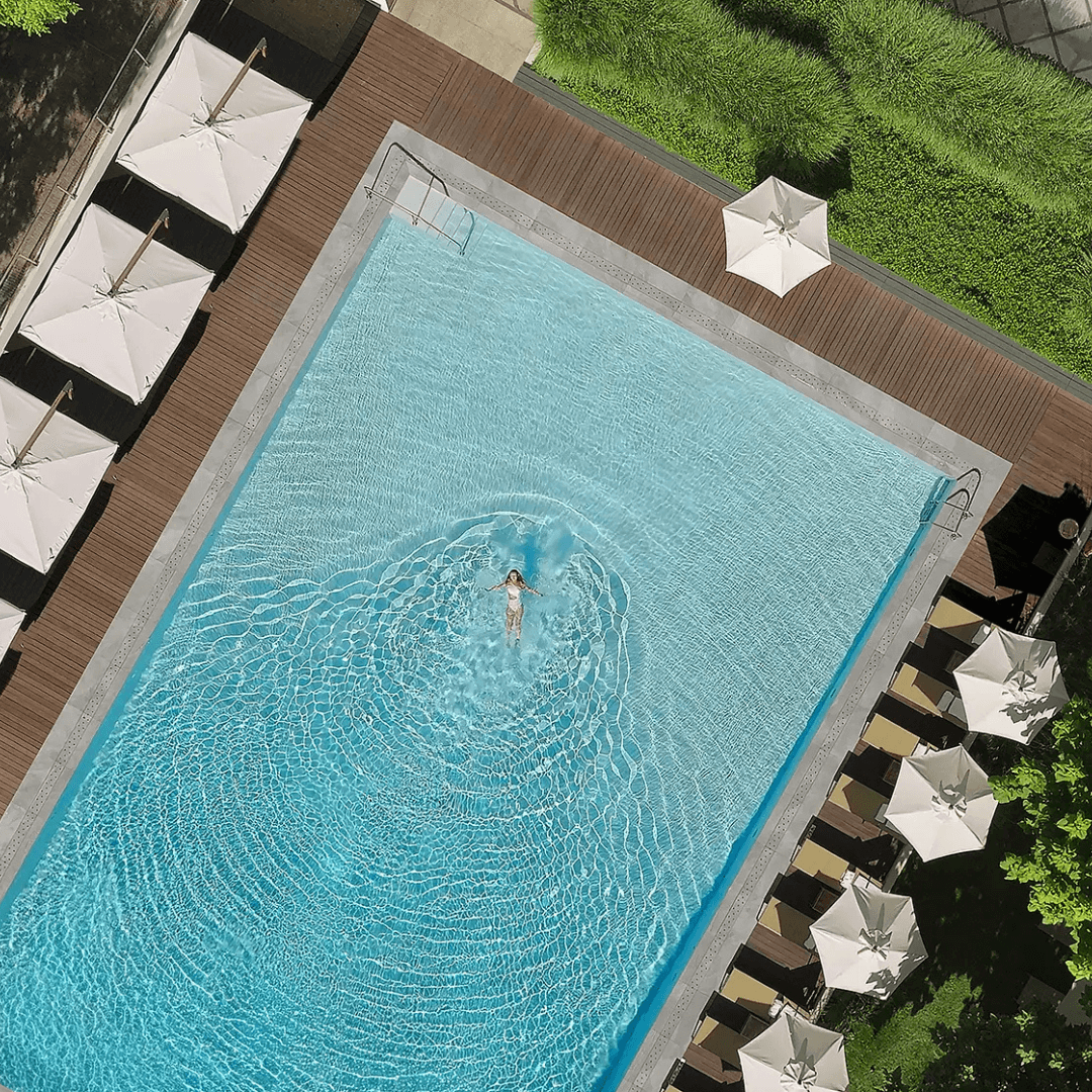 An aerial view of a swimming pool with a person floating in it, surrounded by a deck with white umbrellas and lounge chairs, with greenery nearby.