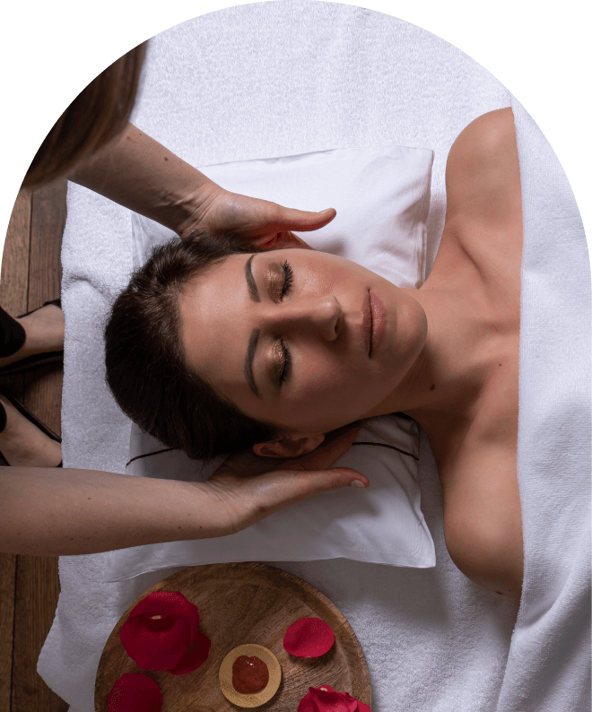 Woman receiving a relaxing head massage in a spa setting, lying on a white towel with red rose petals and a small dish with a candle and rose petals nearby.