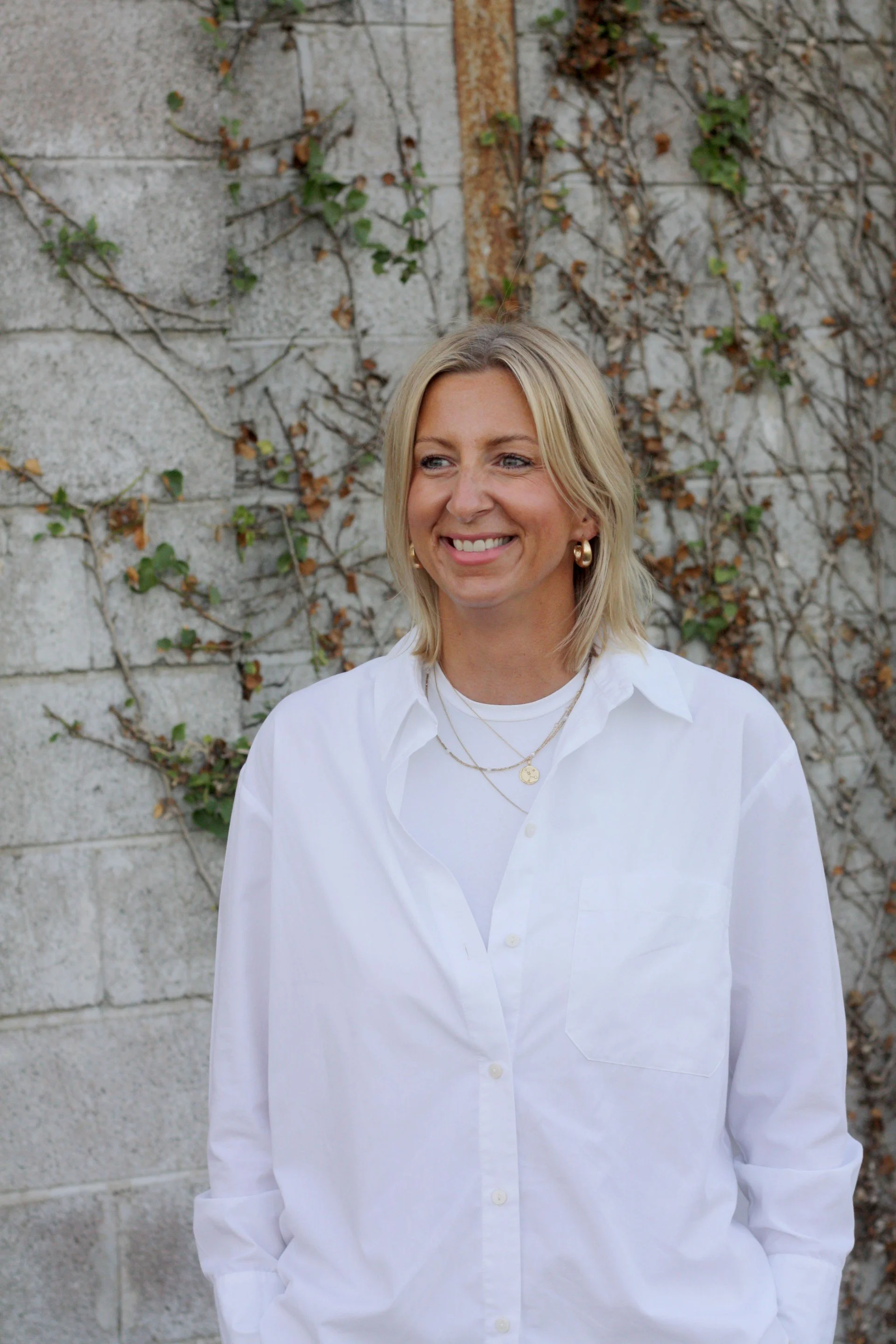 A woman with blonde hair, wearing a white shirt and gold jewelry, standing in front of a concrete wall with ivy growing on it, smiling.