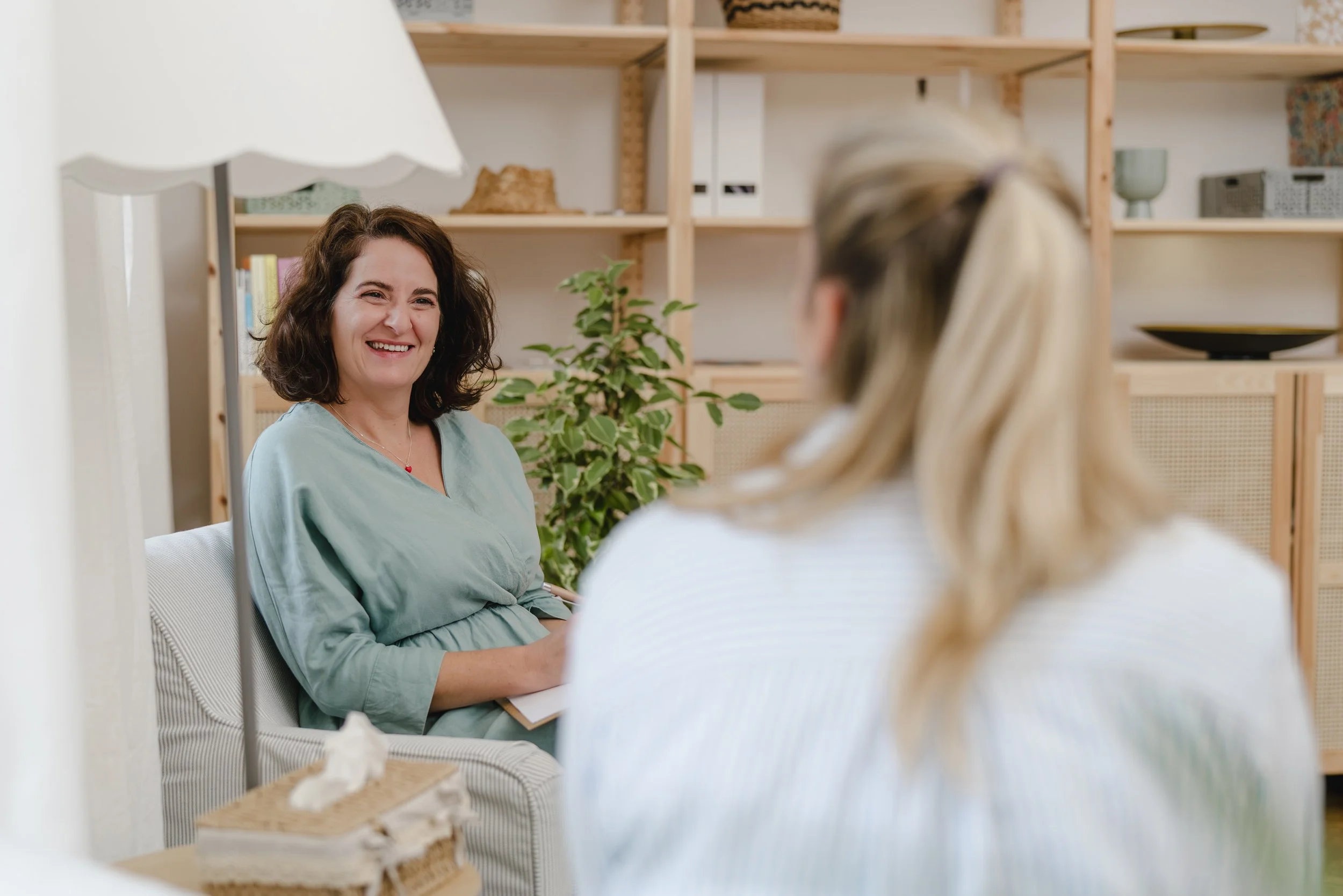 Zwei Frauen in einem Gespräch in einem gemütlichen Raum, eine lacht, die andere hat blonde Haare, im Hintergrund Bücherregal und Pflanzen.