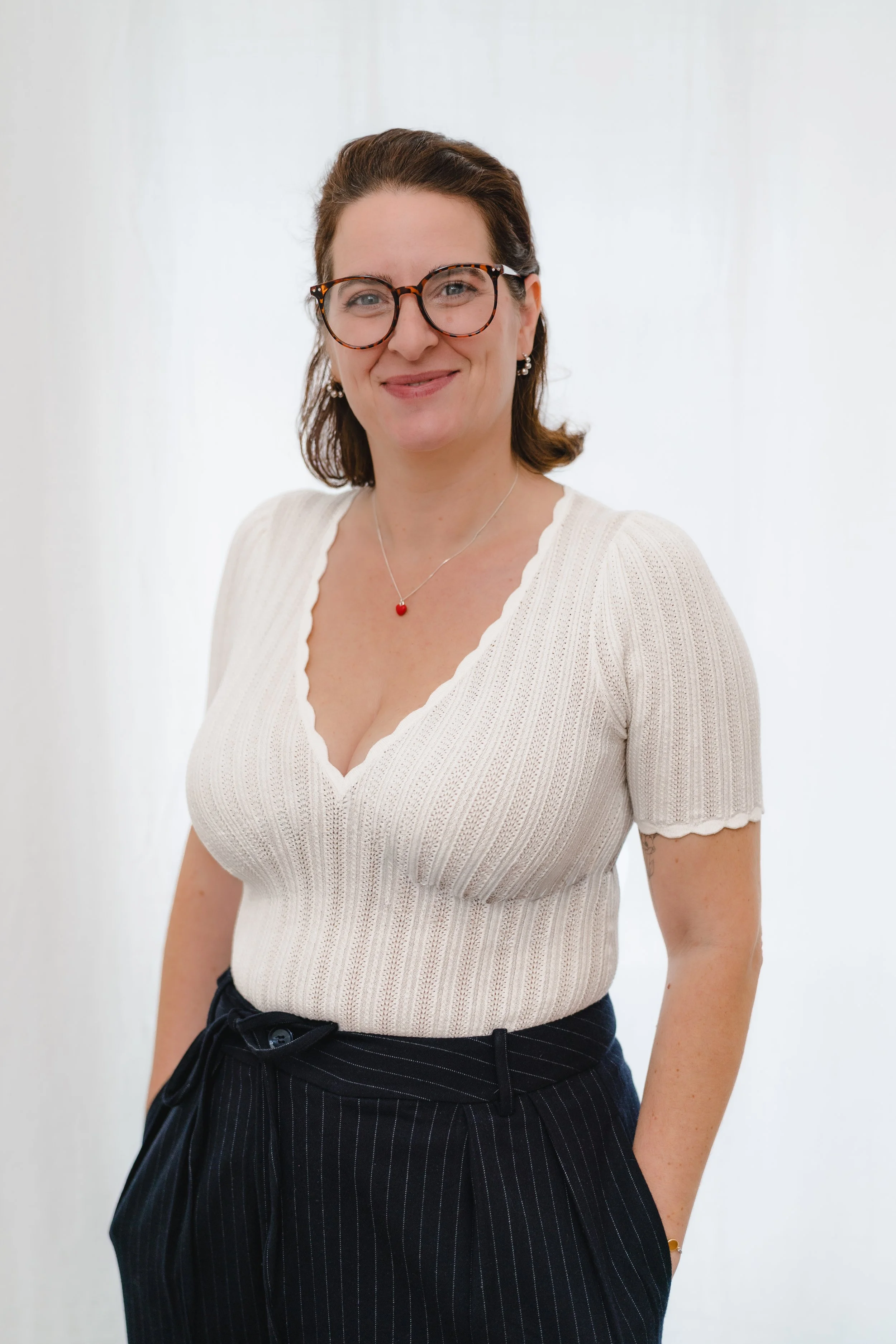 Eine Frau trägt eine weiße, kurze Ärmel, gerippte Bluse mit V-Ausschnitt und schwarze Hosen mit Nadelstreifen, sie lächelt und trägt Brille, Ohrringe und eine Halskette mit rotem Anhänger.