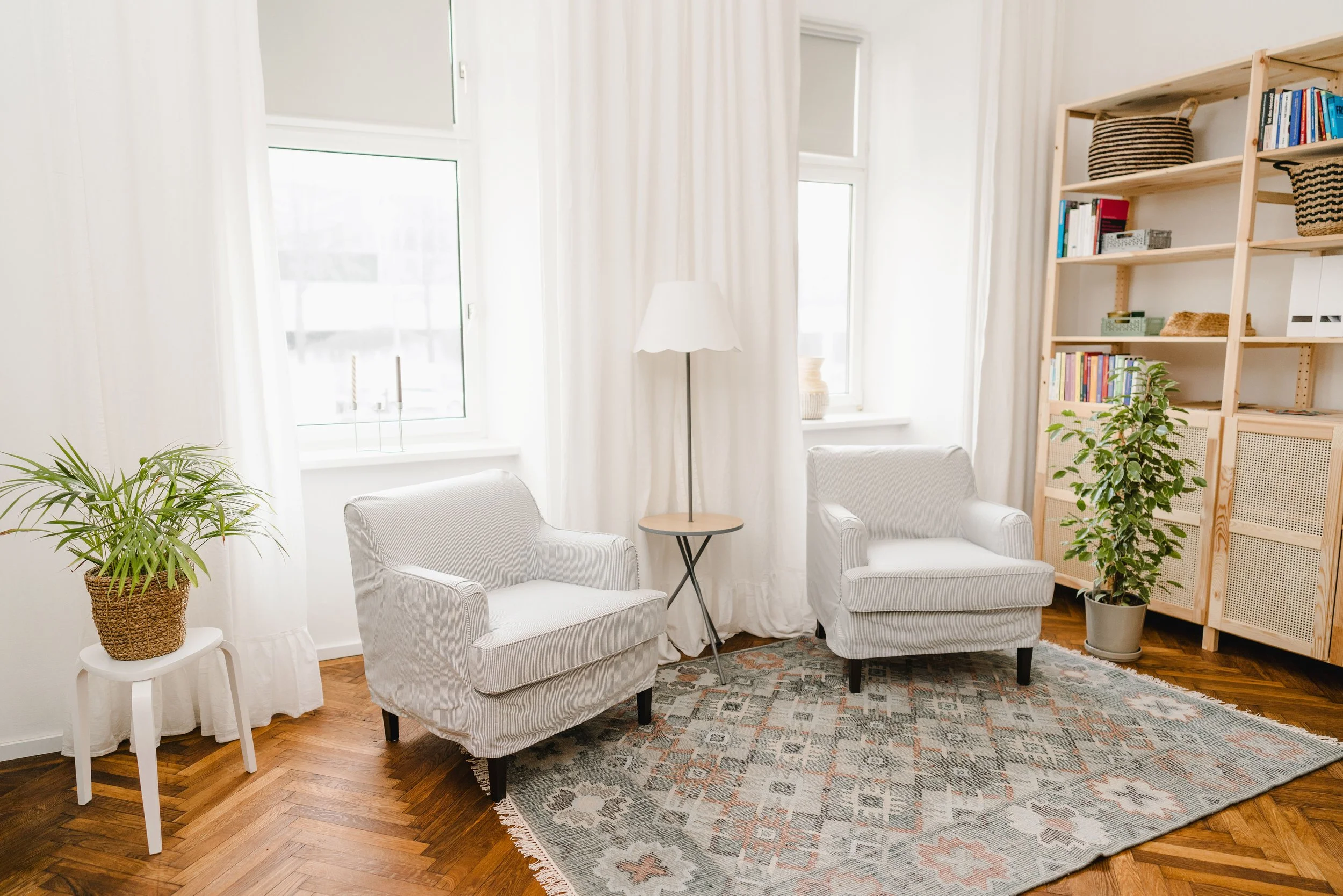 Hell erleuchtetes Wohnzimmer mit zwei weißen Sessel, einem Beistelltisch und Stehlampe, mehrere Fenster mit weißen Vorhängen, ein kleiner brauner Teppich auf dem Holzparkett, eine Stehlampe und ein Bücherregal mit Pflanzen, Büchern und Dekorationsgeg