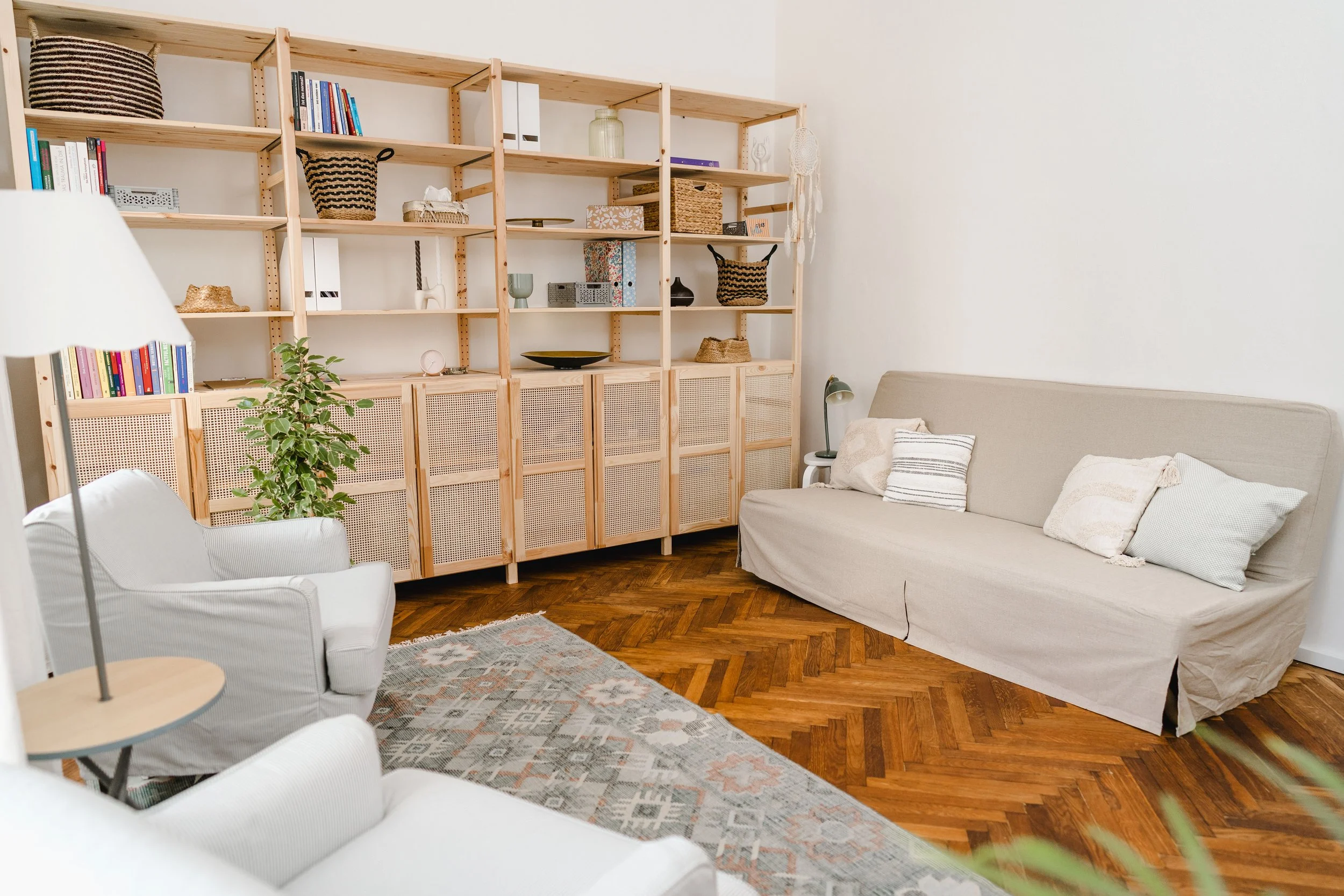 Gemütliches Wohnzimmer mit weißen Sesseln, einem beige Couch mit Kissen, einem Teppich und einem Holzwandregal mit Dekorationsgegenständen und Büchern.