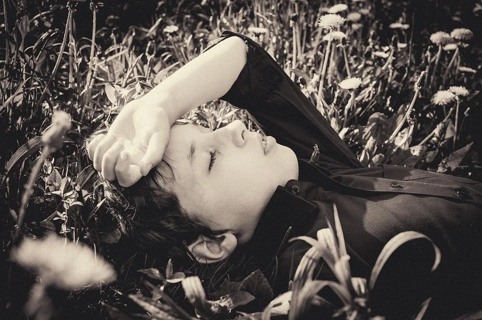 Ein junger Mensch liegt in einem Grasfeld mit closed eyes, mit der Hand auf der Stirn, in schwarz-weiß fotografiert.