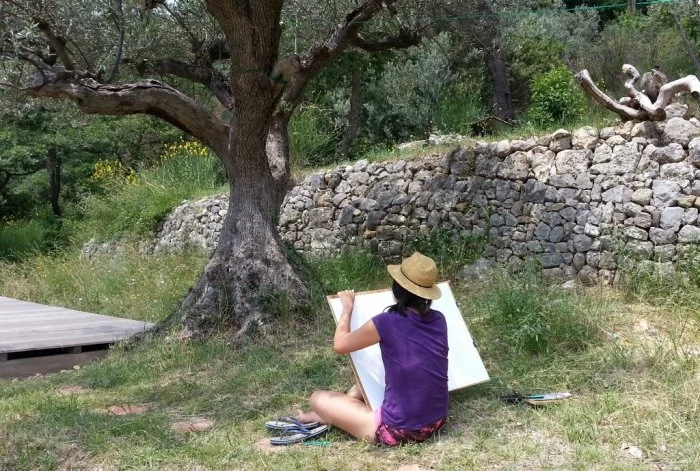 A person wearing a purple top and a wide-brimmed hat sitting on the grass, drawing on a large pad with trees, a stone wall, and greenery in the background.