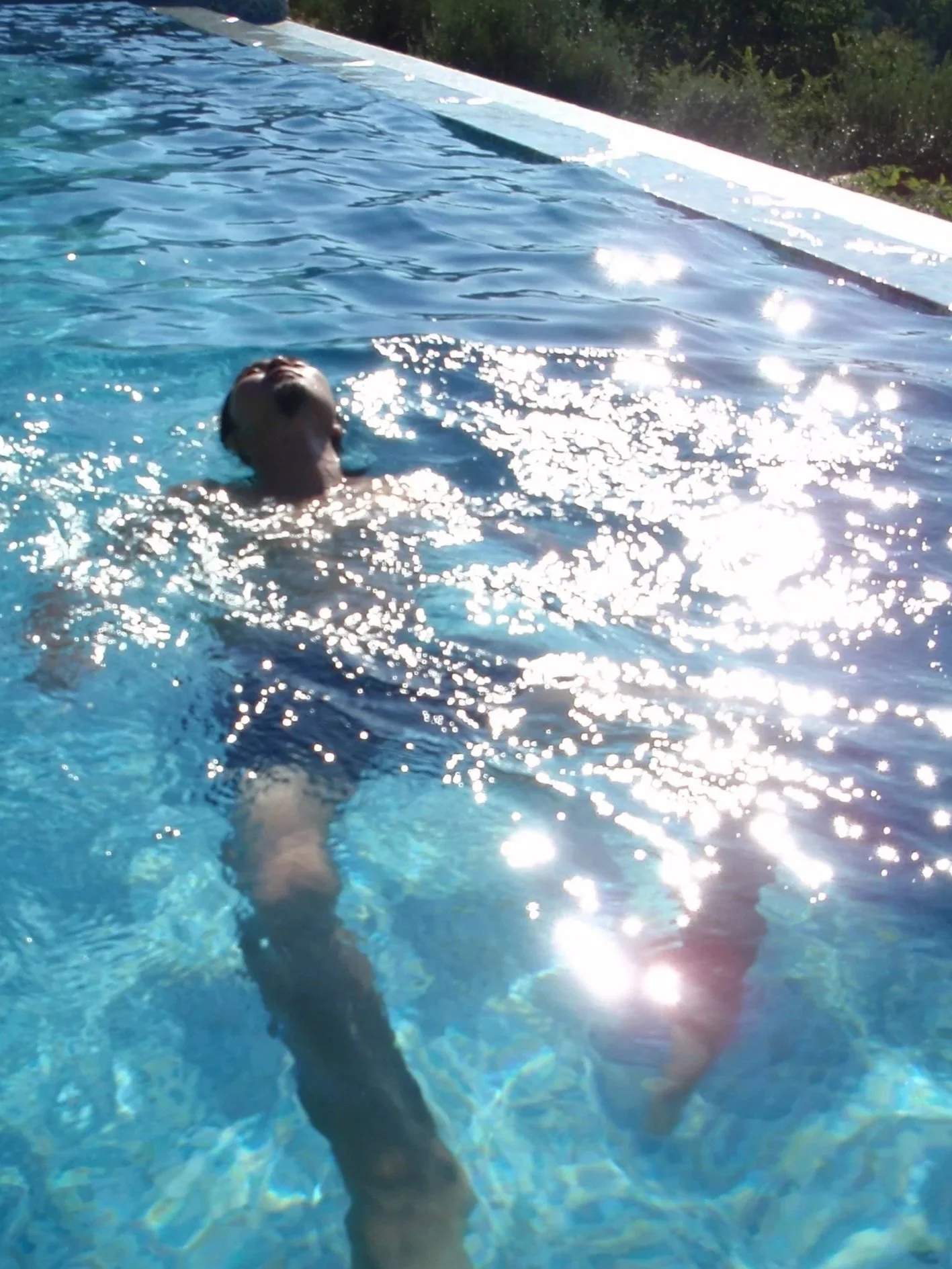 A person swimming in a pool on a sunny day, with water shimmering in sunlight and a white pool edge visible at the top of the image.