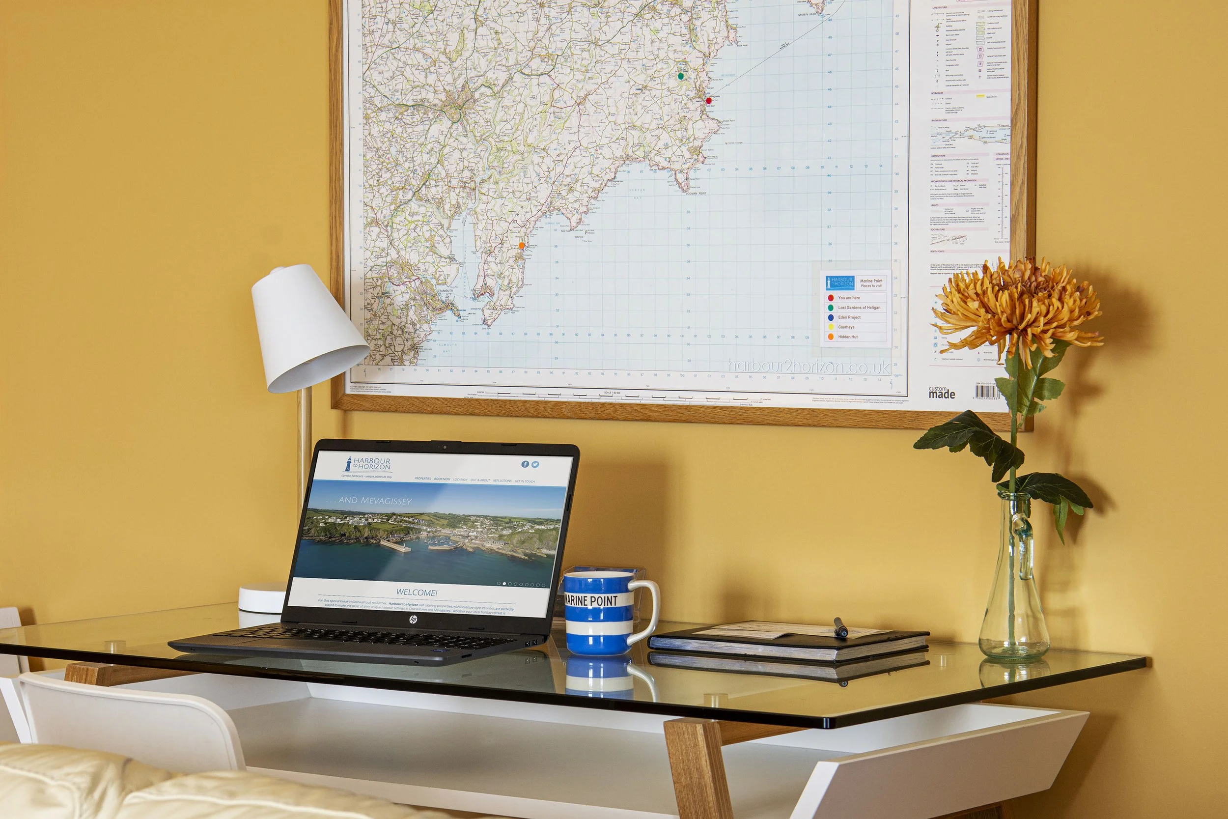 Close up of the desk in Marine Point, with laptop and coffee mug. The wall behind is a warm yellow with a framed map of the local area hanging above.