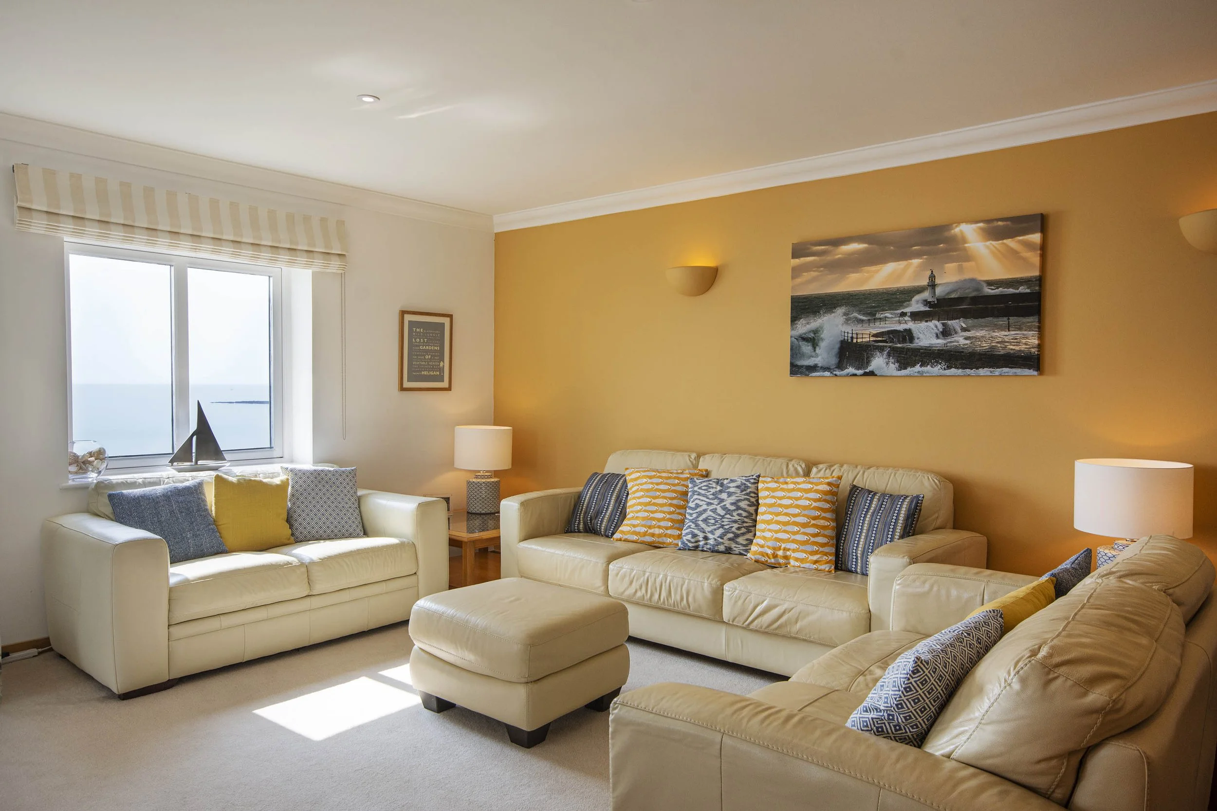 Comfy sofas and cushions in the living room of Marine Point, warm colours of yellows and creams. View out of a smaller window of the sea in the background.