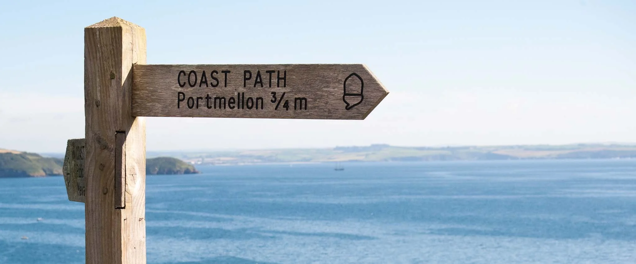 Close up of a wooden coast path arrow sign with the words 'Portmellon 3/4 mile' engraved on it. The sea and coastline are in the background.