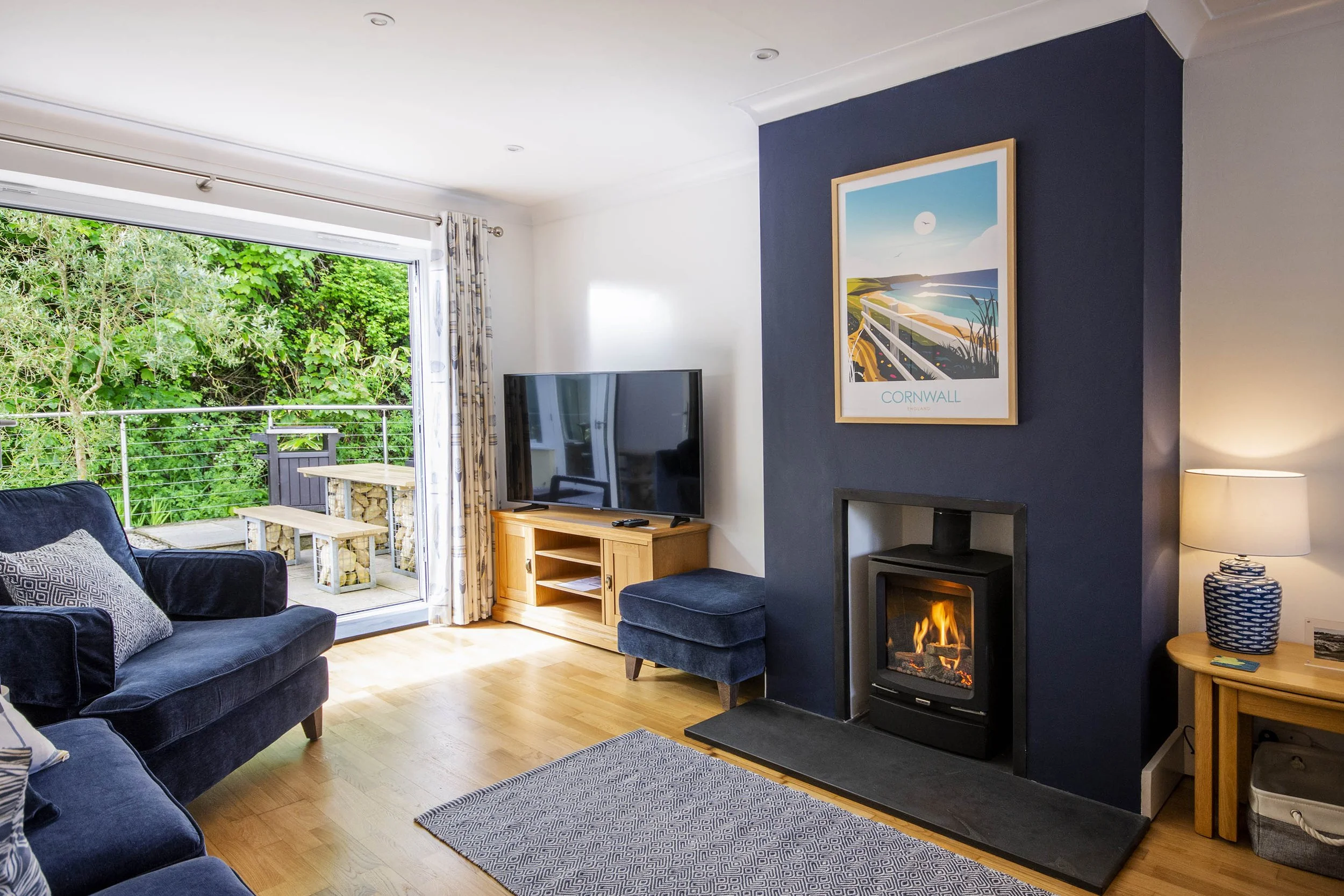 Interior view of the living room at Tall Ships, with comfy sofas, log burner, wooden floors and a rug. There are art prints on the walls, and the patio doors are opened on to the terrace.
