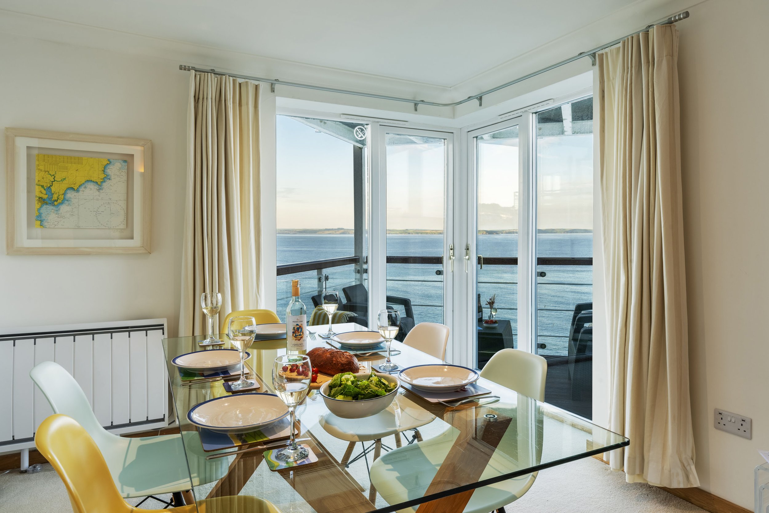 Interior view of the living room at the Marine Point apartment, a table is in the foreground set for lunch and there's a view out of the balcony windows in the background looking over the fishing port of Mevagissey
