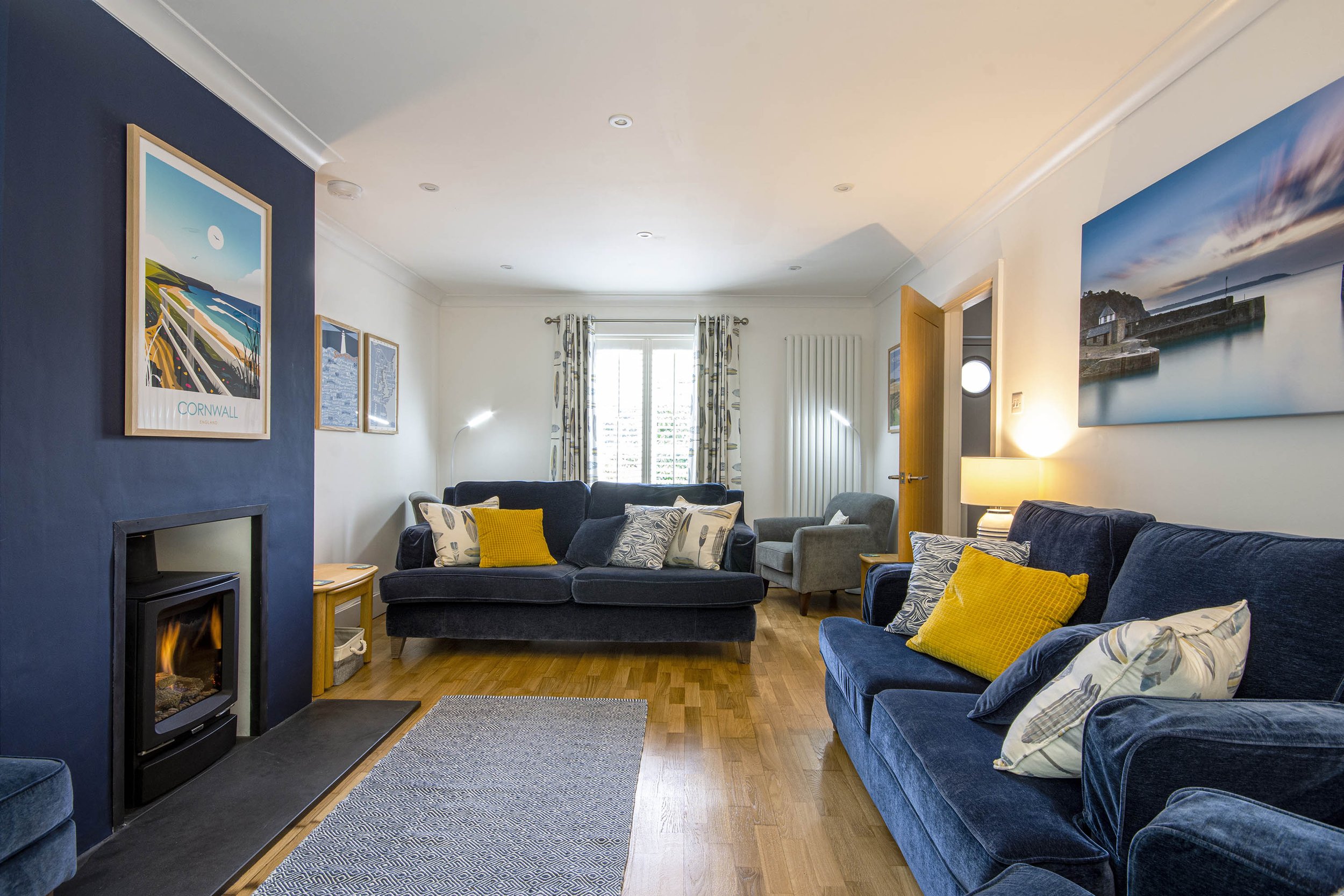 Interior view of the living room at Tall Ships, with comfy dark blue sofas, log burner, wooden floors and a rug. There are art prints on the walls.