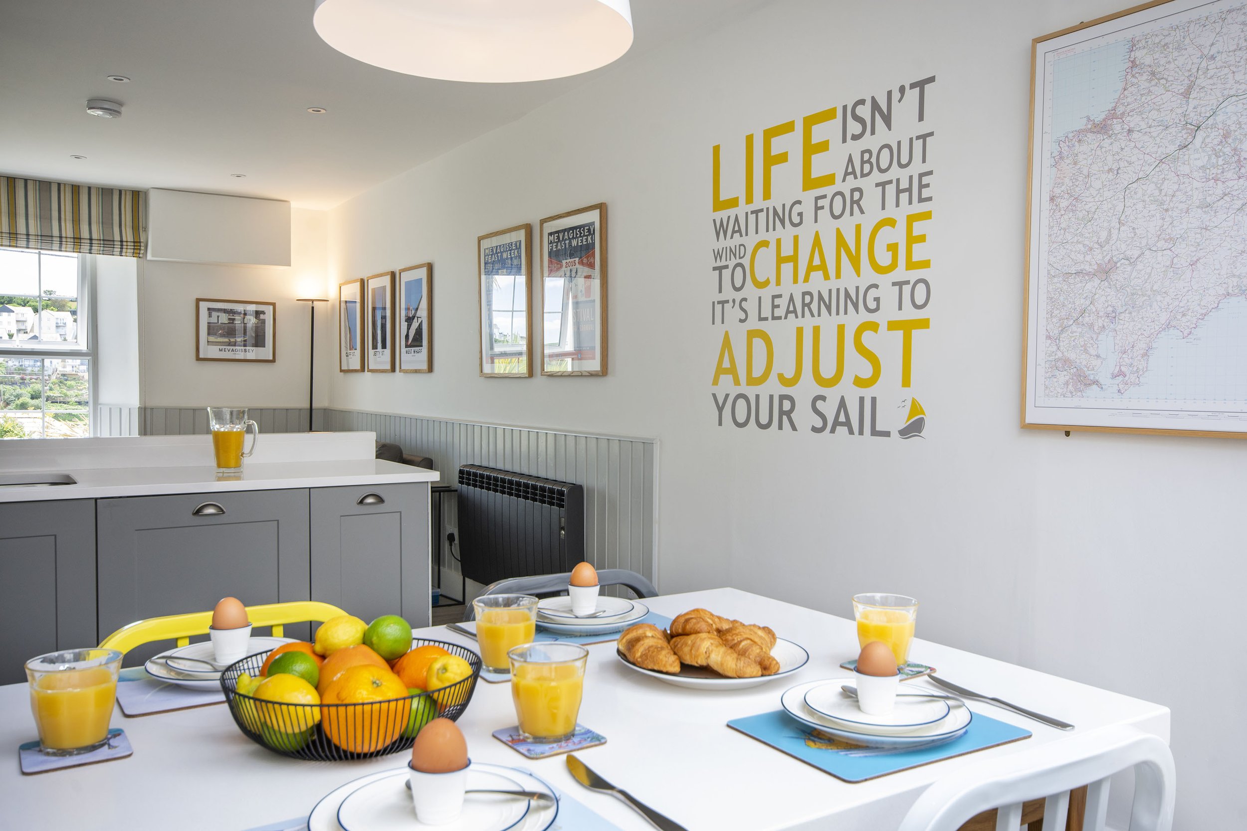 Interior view of the kitchen diner in Seafarers cottage. There is colour art on the walls and the table is set for breakfast. The living room is in the background.