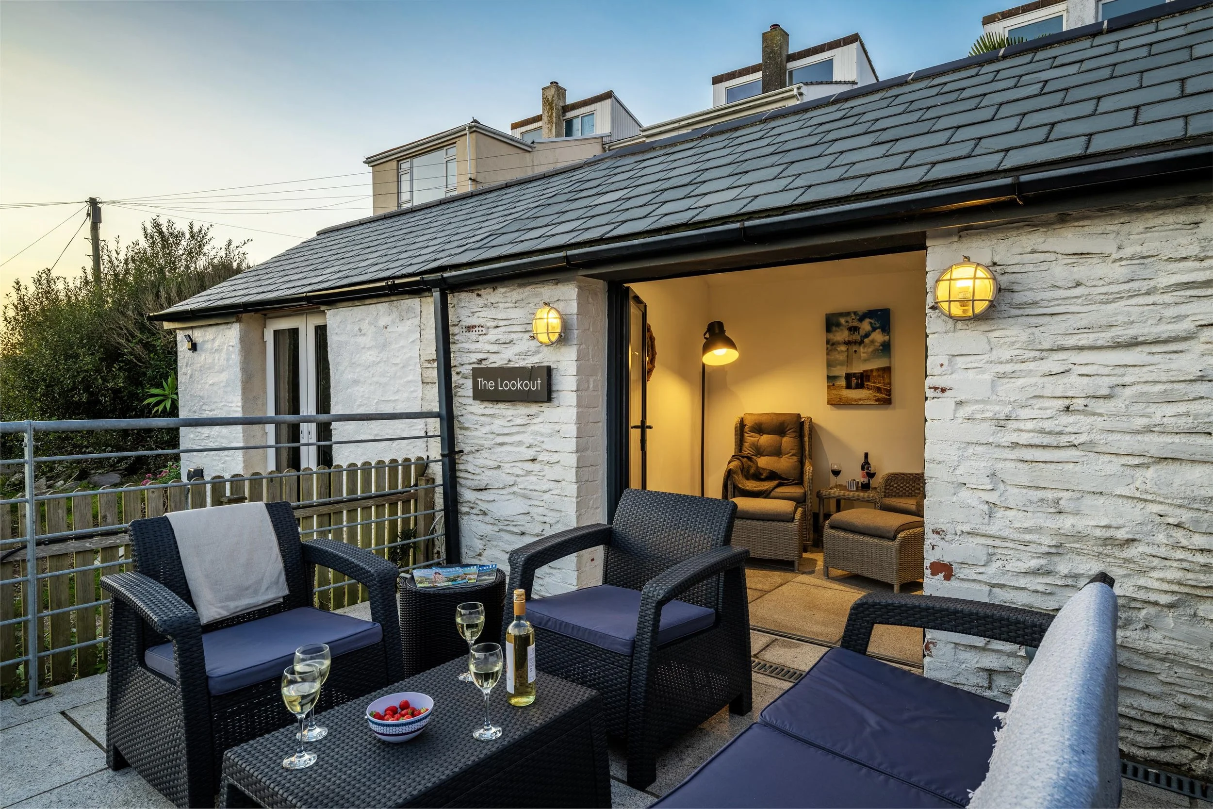 Terrace and patio of Seafarers cottage with garden room at the back with interior seating. It is evening and the outside area is lit subtly. There are garden chairs and sofas outside on the patio. 