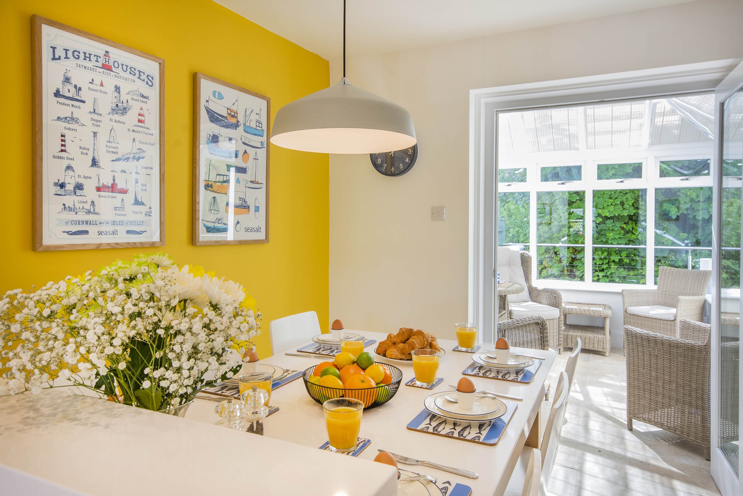 Interior view of the kitchen and diner at Tall Ships. The wall behind the dining table is painted yellow with bright art prints hanging up. The table is set for breakfast and there are flowers on the counter top. The doors are open in to the conserva