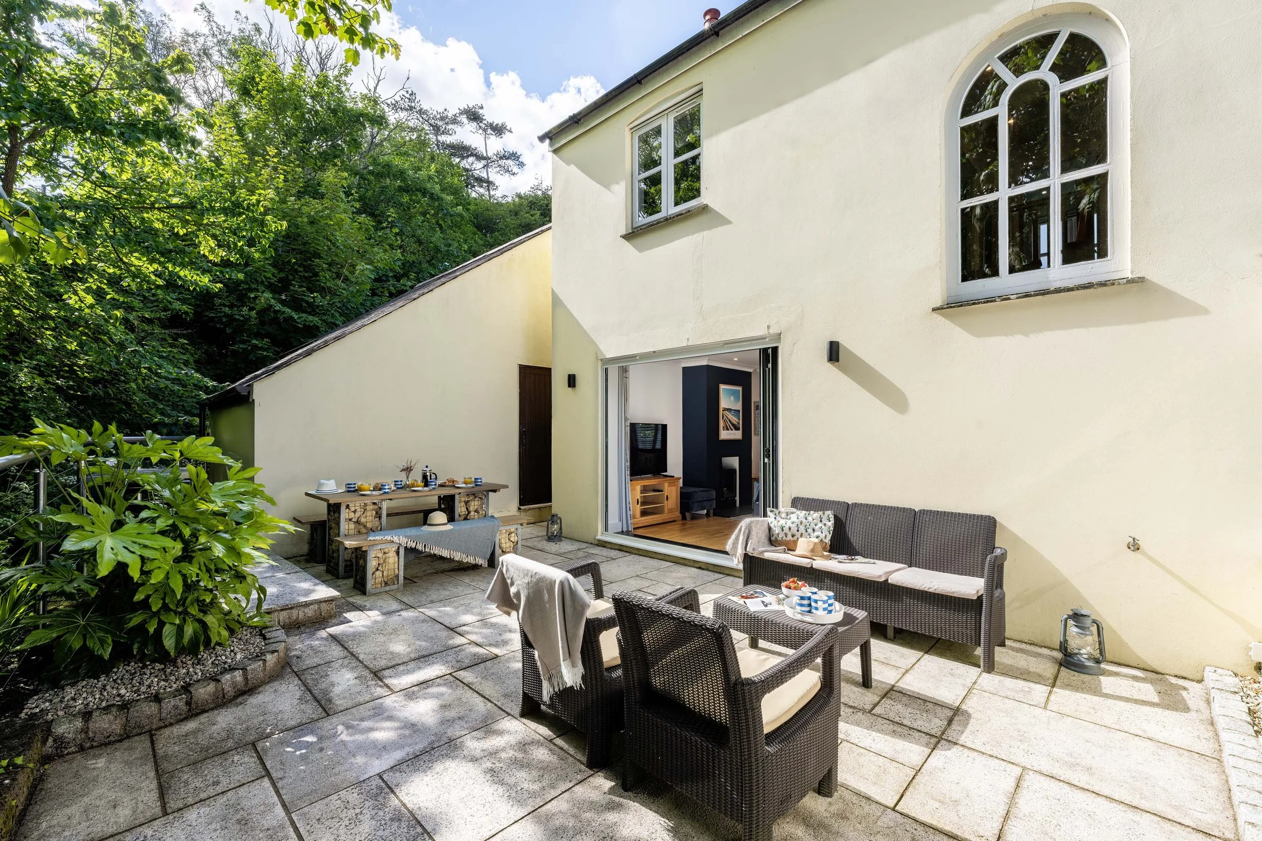 Day time view of the patio to the rear of the Tall Ships property with picnic table and patio furniture with cushions and throws. There are trees in the background.