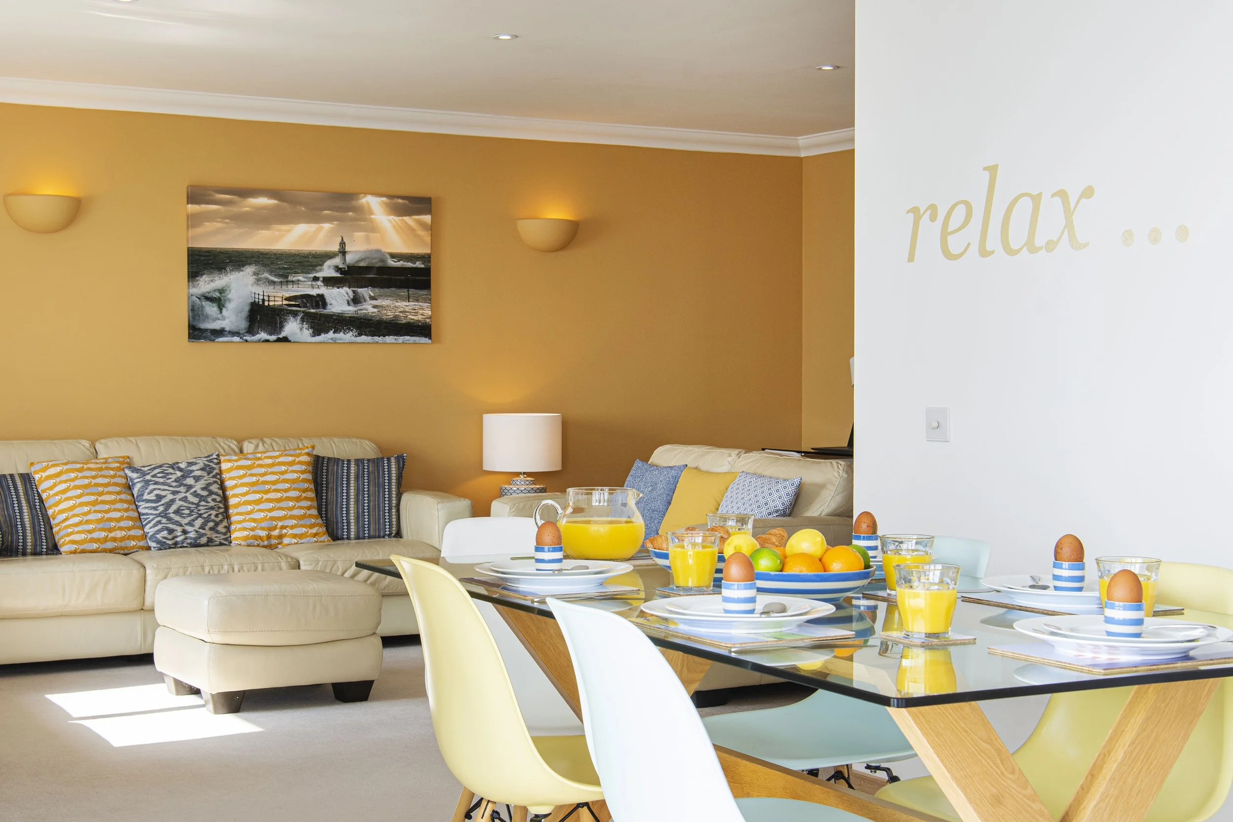 Interior view of the Marine Point apartment, with dining table set for breakfast in the foreground. Comfy sofas and cushions are in the living room in the background, warm colours of yellows and creams. 