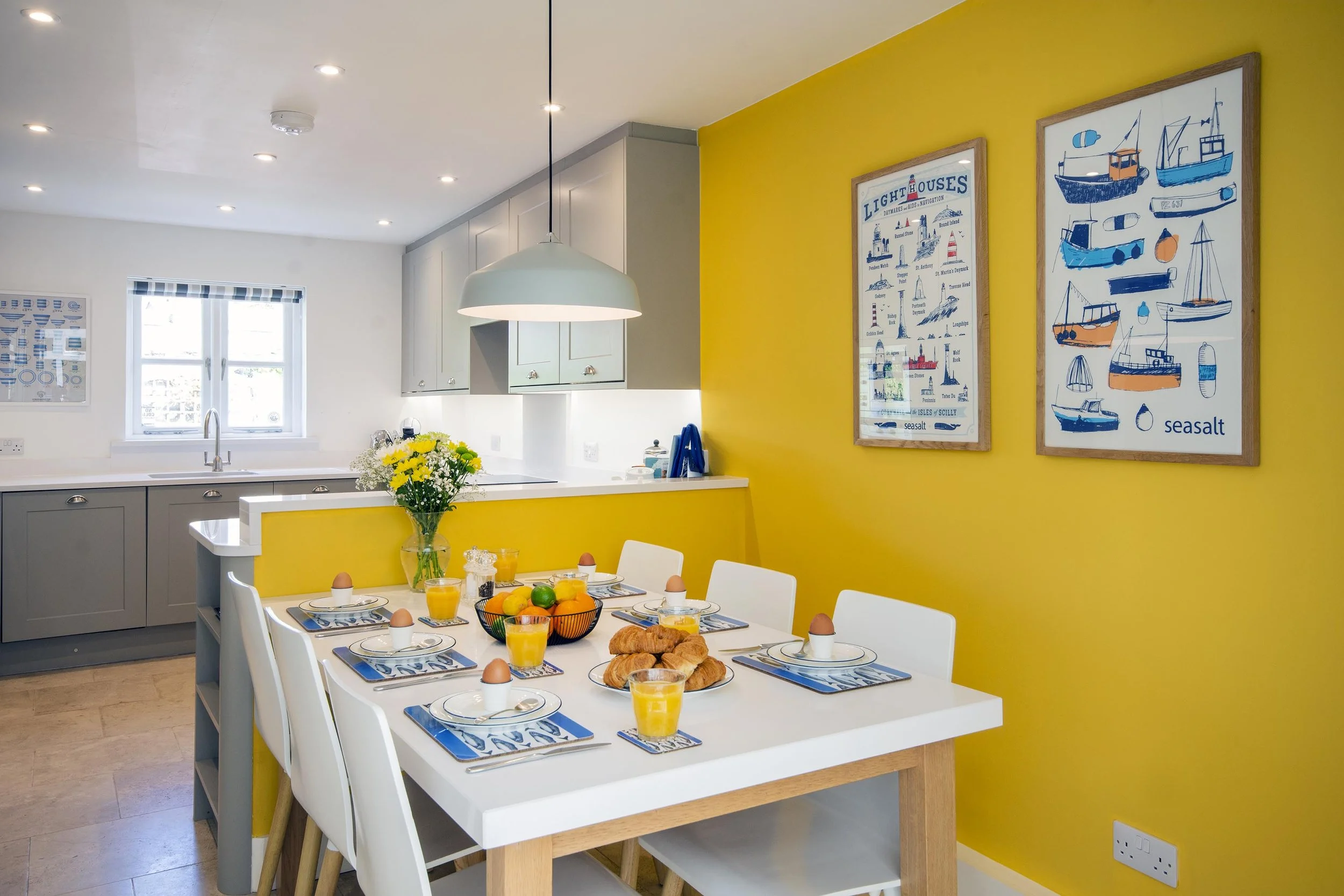 Interior view of the kitchen and diner at Tall Ships. The wall behind the dining table is painted yellow with bright art prints hanging up. The table is set for breakfast and there are flowers on the counter top.