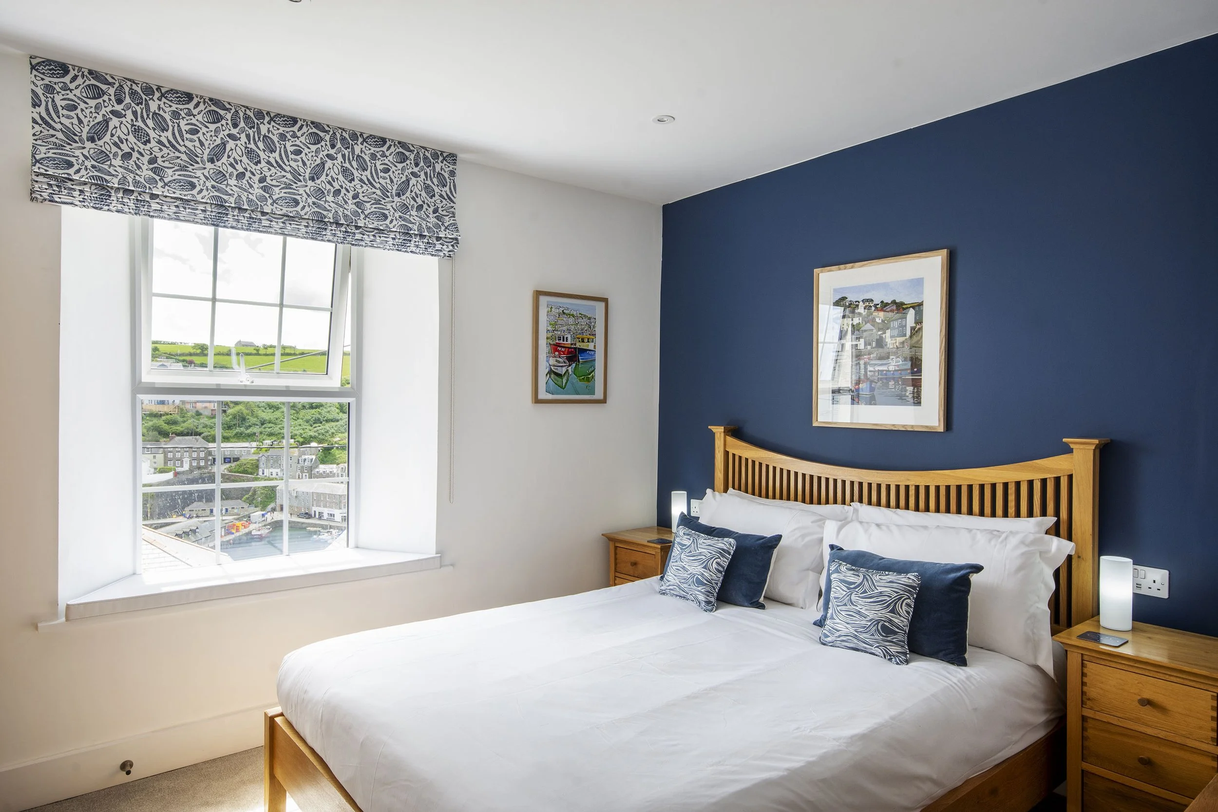 Main bedroom at Seafarers, with large double bed and the wall behind it painted dark blue. There is a view out of the window overlooking Mevagissey harbour.