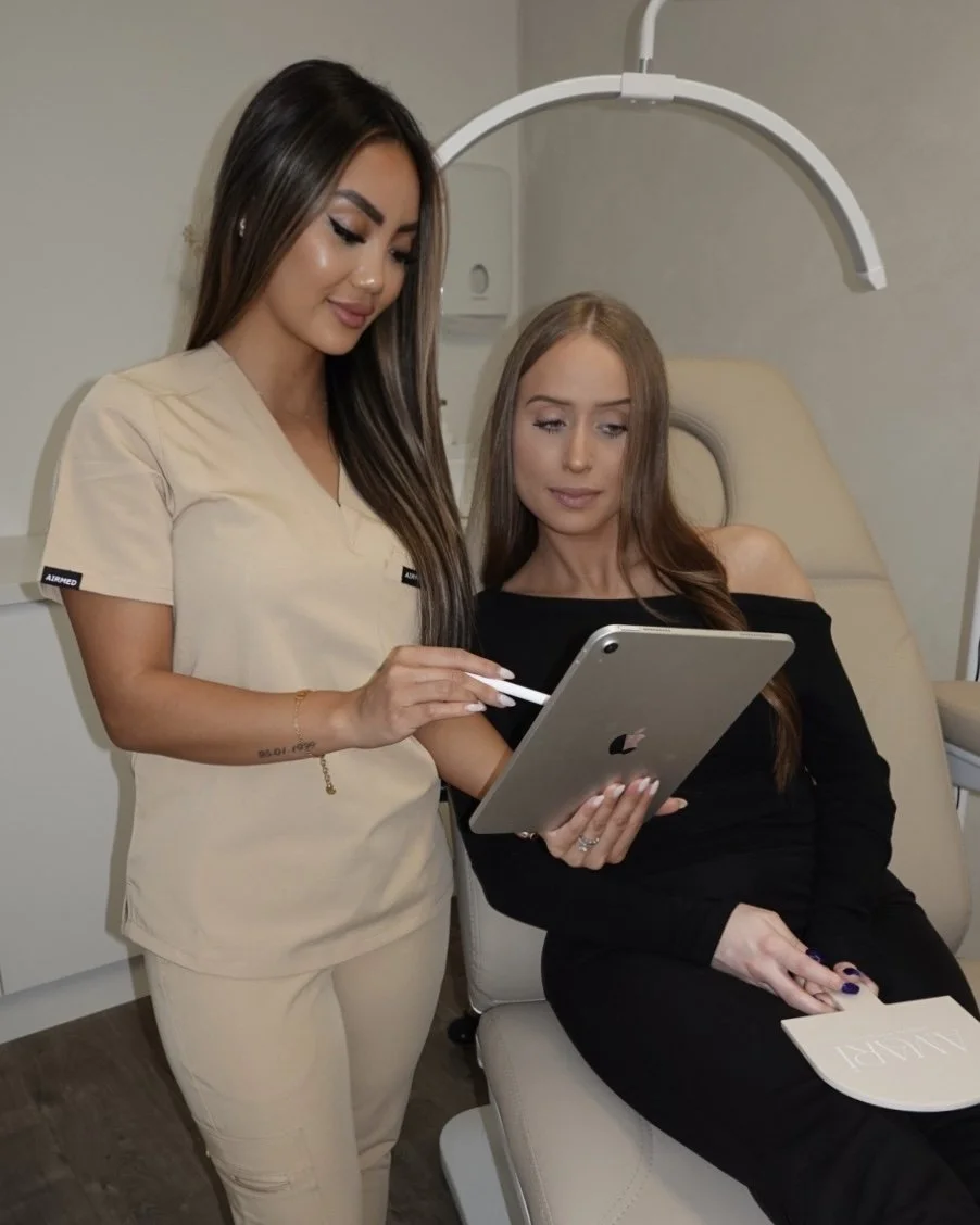 A woman in beige scrubs shows an iPad to a woman seated in a medical chair in a clinical setting.
