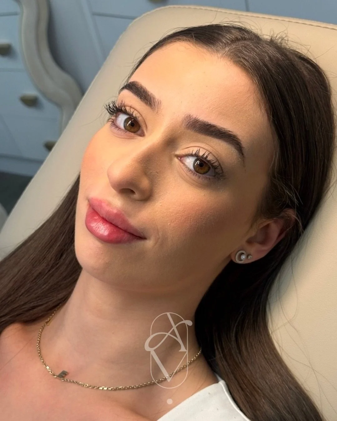 A woman with brown hair lying on a beige chair, wearing earrings and a necklace, looking at the camera with a slight smile.