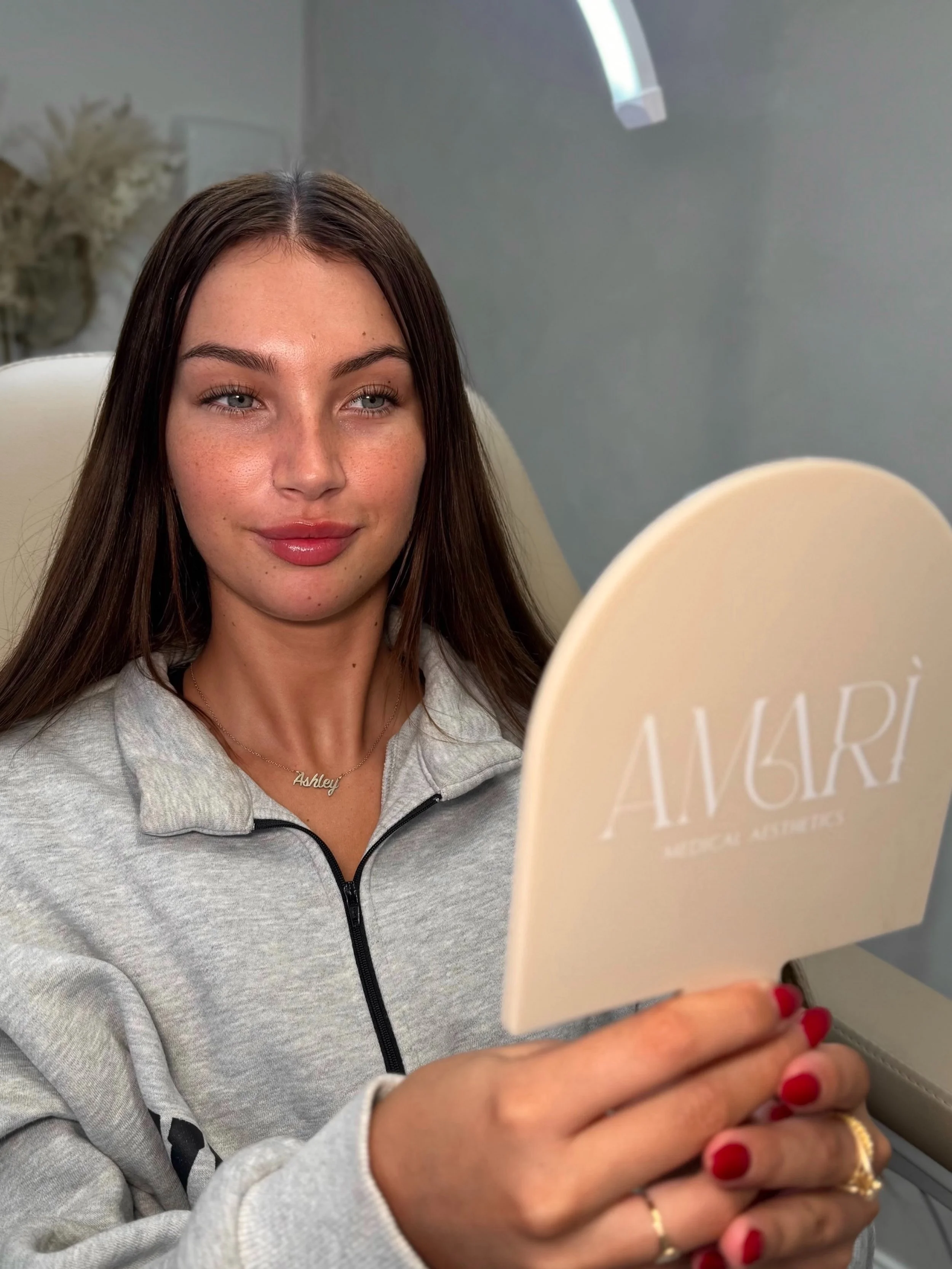 A young woman with long brown hair and blue eyes examining her face in a mirror, wearing a gray zip-up hoodie, with red nail polish, gold rings, a delicate necklace with the name Ashley, and sitting in a skincare or aesthetic clinic.