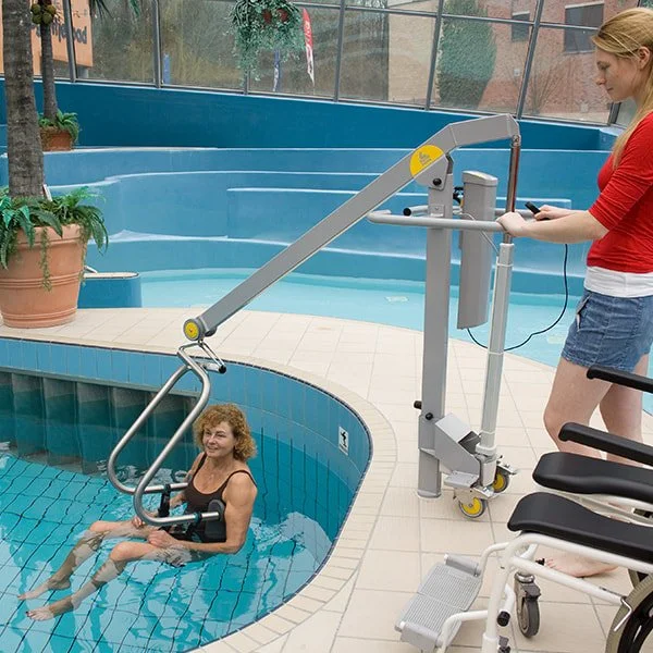 A woman in a small indoor pool lifting her leg with aquatic therapy equipment, assisted by a woman standing outside the pool, in a therapy or rehabilitation setting.
