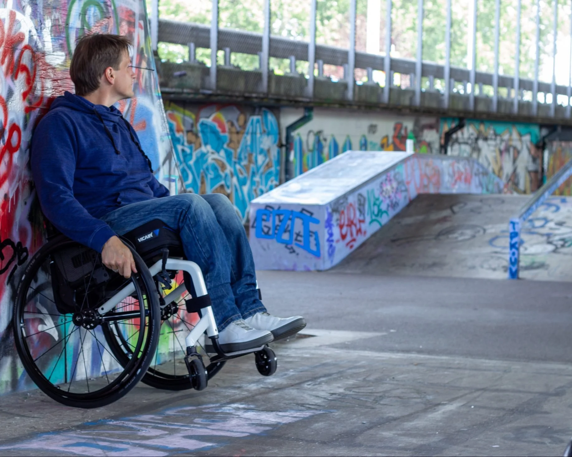 A young man in a wheelchair sitting at a graffiti-covered skate park wall, looking to the side, wearing a blue hoodie and jeans.