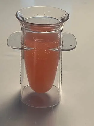 A glass cup filled with a pinkish liquid, placed on a clear plastic stand.
