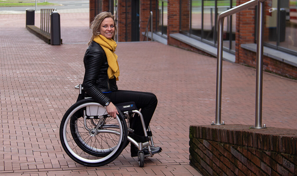 A woman in a wheelchair on a brick sidewalk outside a building, smiling at the camera.