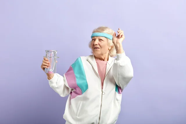 An elderly woman in exercise attire holding a beaker and wearing a headband, standing against a light purple background.
