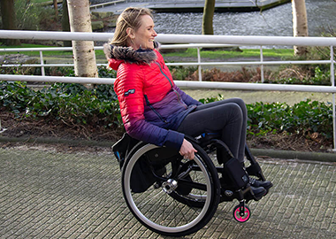 Woman in a red and purple jacket sitting in a wheelchair on a paved path along a river