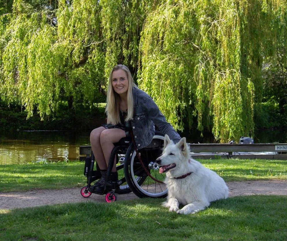 A young woman in a wheelchair outdoors next to a white dog, with a lush green tree and a body of water in the background.