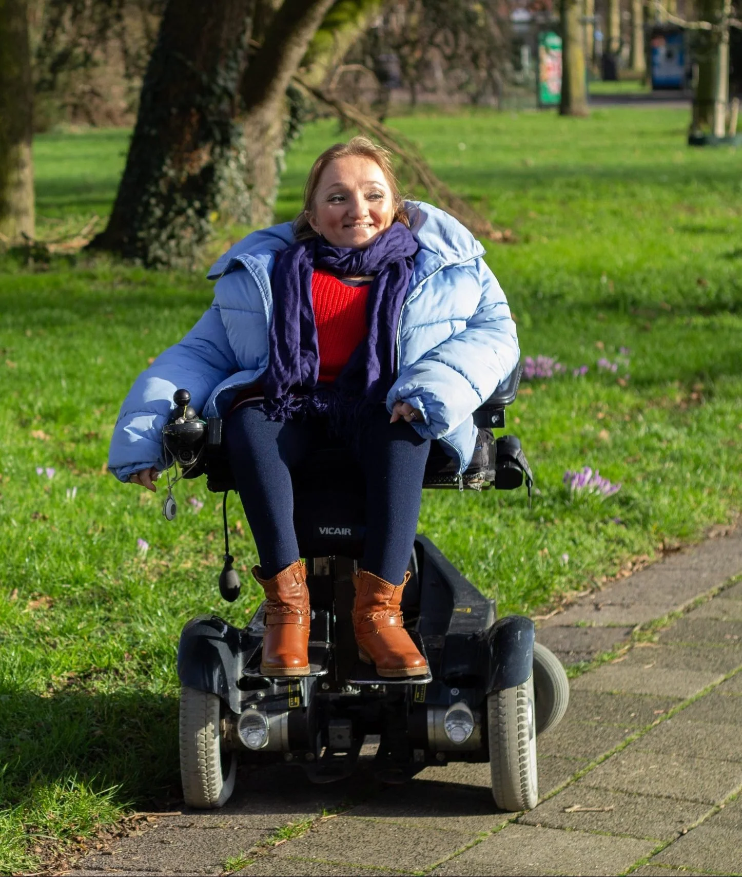 A smiling woman in a wheelchair outdoors on a grassy park path, wearing a blue puffy jacket, red sweater, dark pants, brown boots, and a purple scarf.