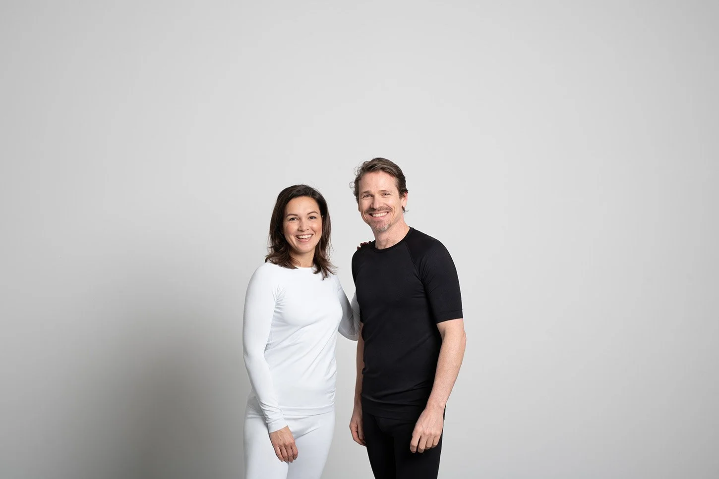 A smiling woman and man standing closely together against a plain white background, with the woman's hand on the man's shoulder.