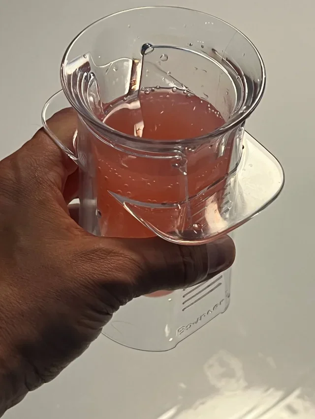 A hand holding a clear measuring cup filled with pinkish liquid, with a transparent plastic product on top, and a white surface below.