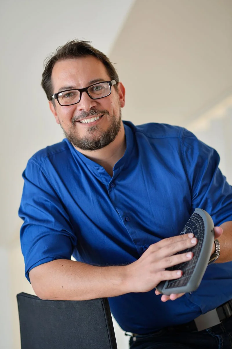 A smiling man with glasses and a beard, wearing a blue shirt, holding a computer keyboard in his right hand while leaning on a chair.