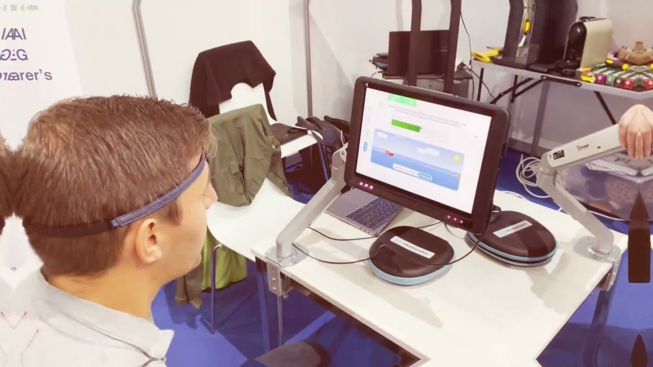 A person wearing a headband using a touchscreen device connected to robotic arms on a table, with a laptop nearby, in a lab or exhibition setting.