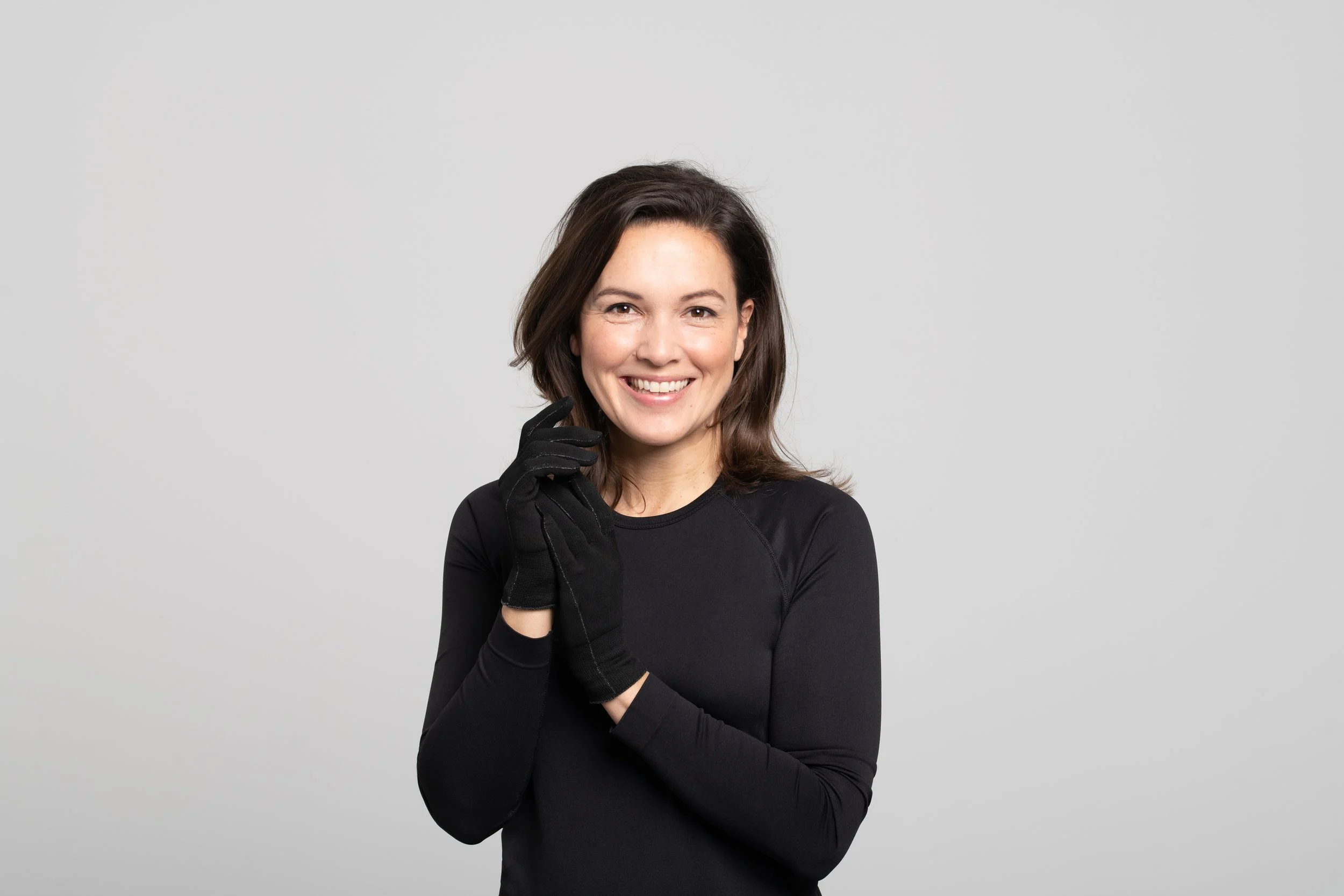 A woman with shoulder-length brown hair smiling, wearing a black long-sleeve shirt and black gloves, standing against a plain light gray background.