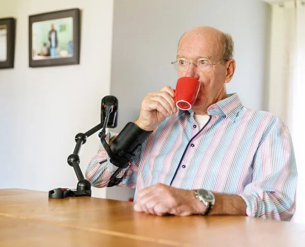 An elderly man with a prosthetic arm is sitting at a table, drinking from a red cup and smiling.