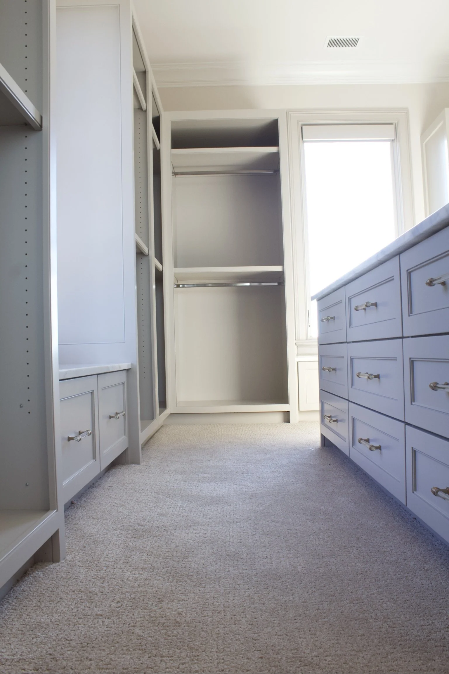 Custom closet cabinets painted Sherwin Williams Anew Gray, featuring stacked drawer island, window seats, hidden closet behind mirror, and natural stone counter tops by Crawford Cabinets